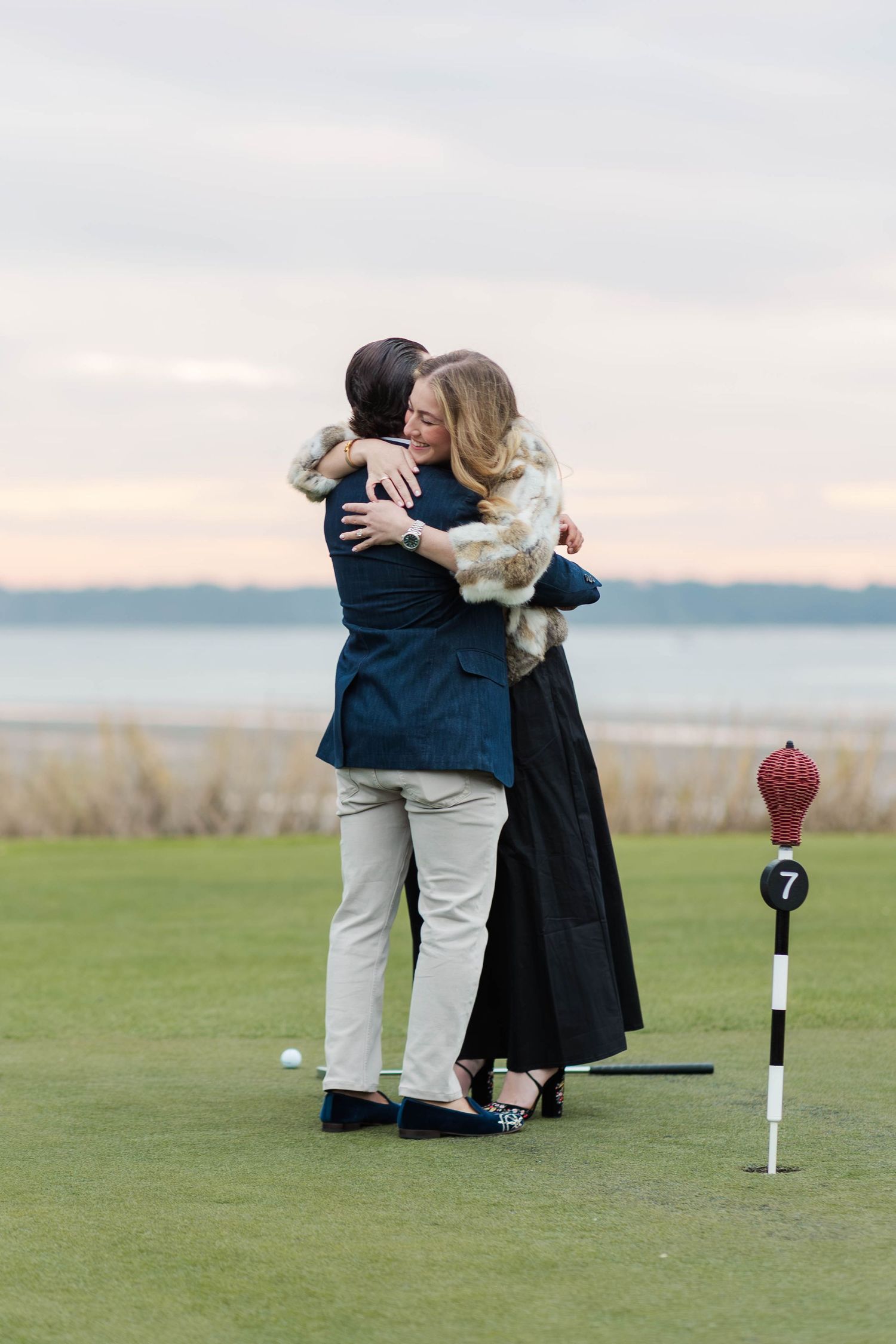 A couple embraces on a golf course during sunset with a scenic coastal view in the background.