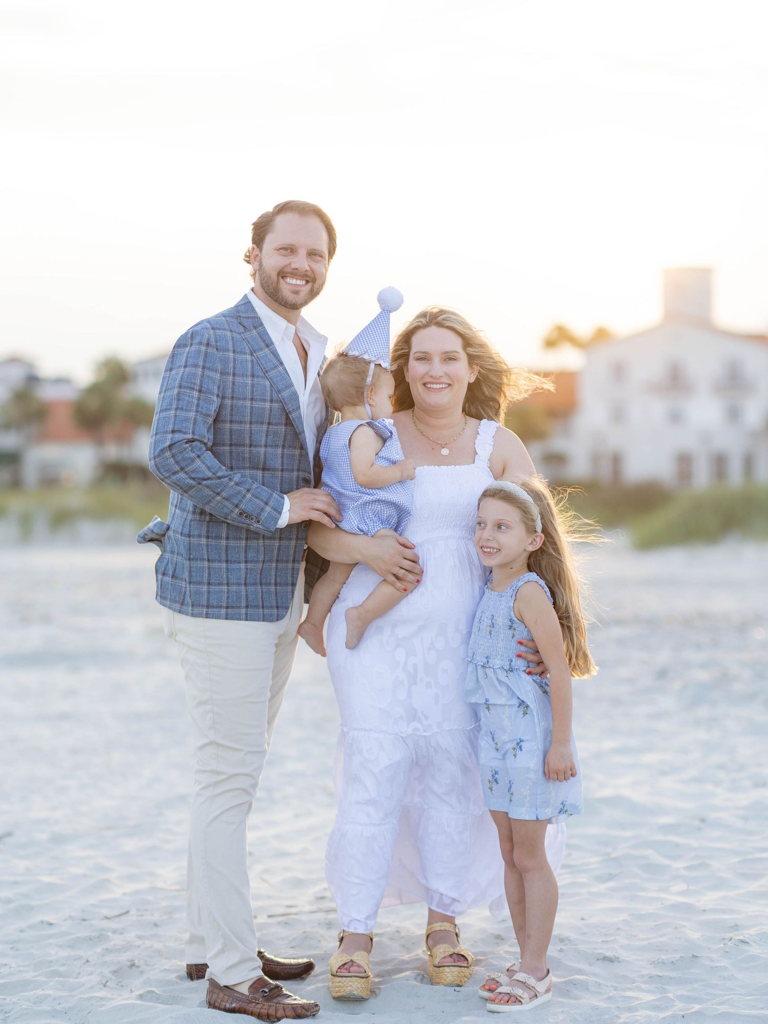Family of four posing on a sandy beach at sunset, dressed in coordinating blue and white outfits.