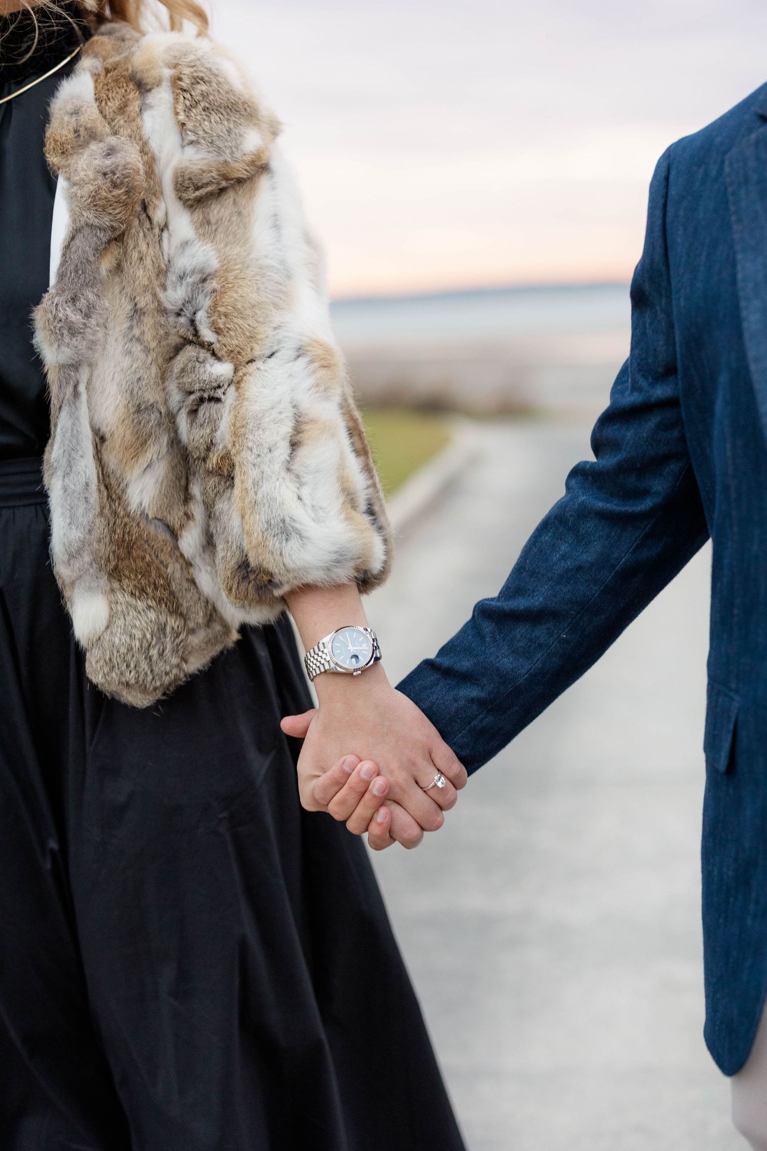 Close-up of couple holding hands, with one wearing a fur vest and black dress, the other in a navy suit at sunset.