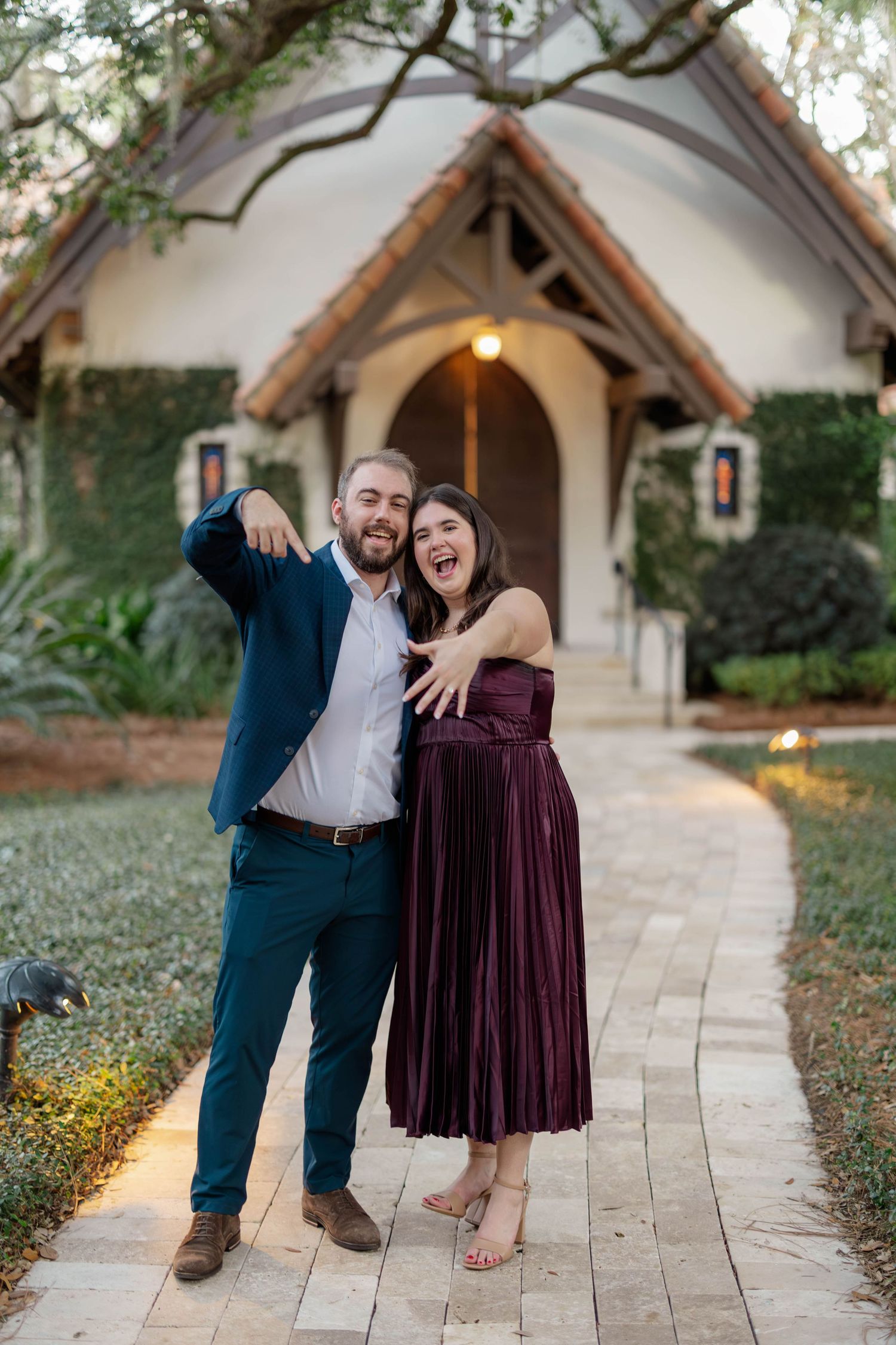 Joyful couple showing off engagement ring outside a charming chapel on a stone pathway at dusk.