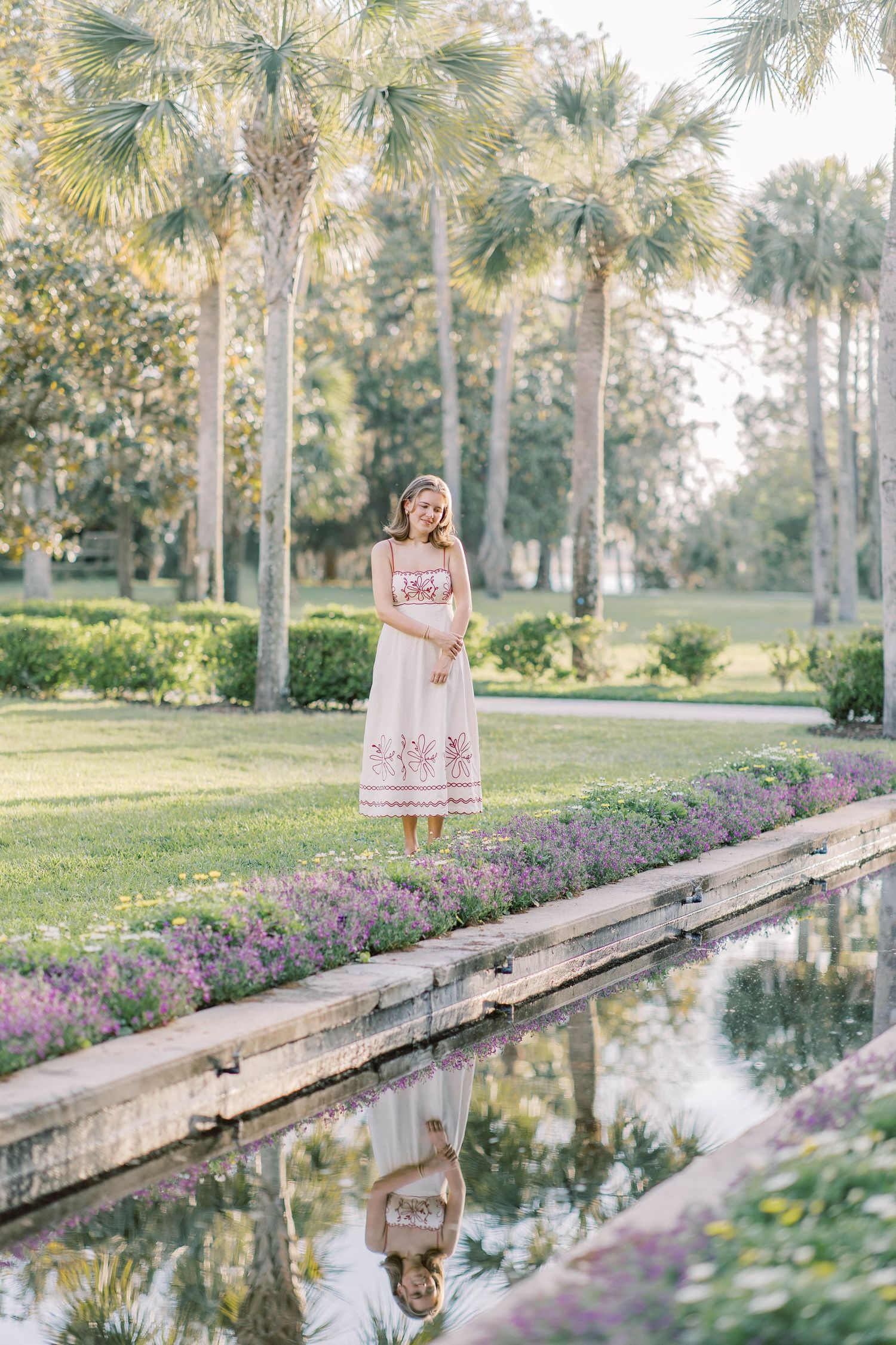 A woman in a white floral dress stands by a reflective garden pond surrounded by palm trees and purple flowers.