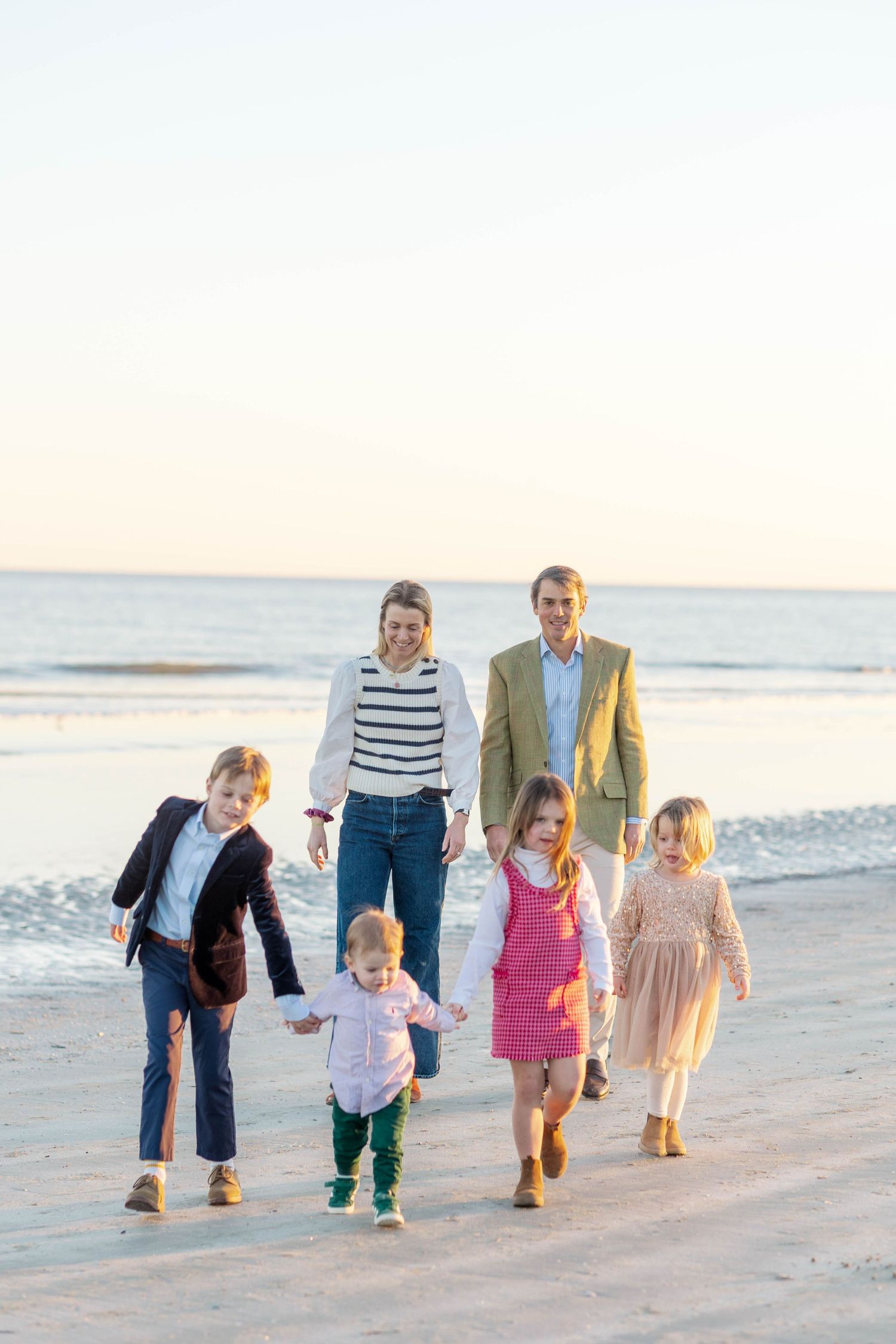 Family of six walking on beach at sunset, two parents and four children in colorful outfits, ocean waves in background.