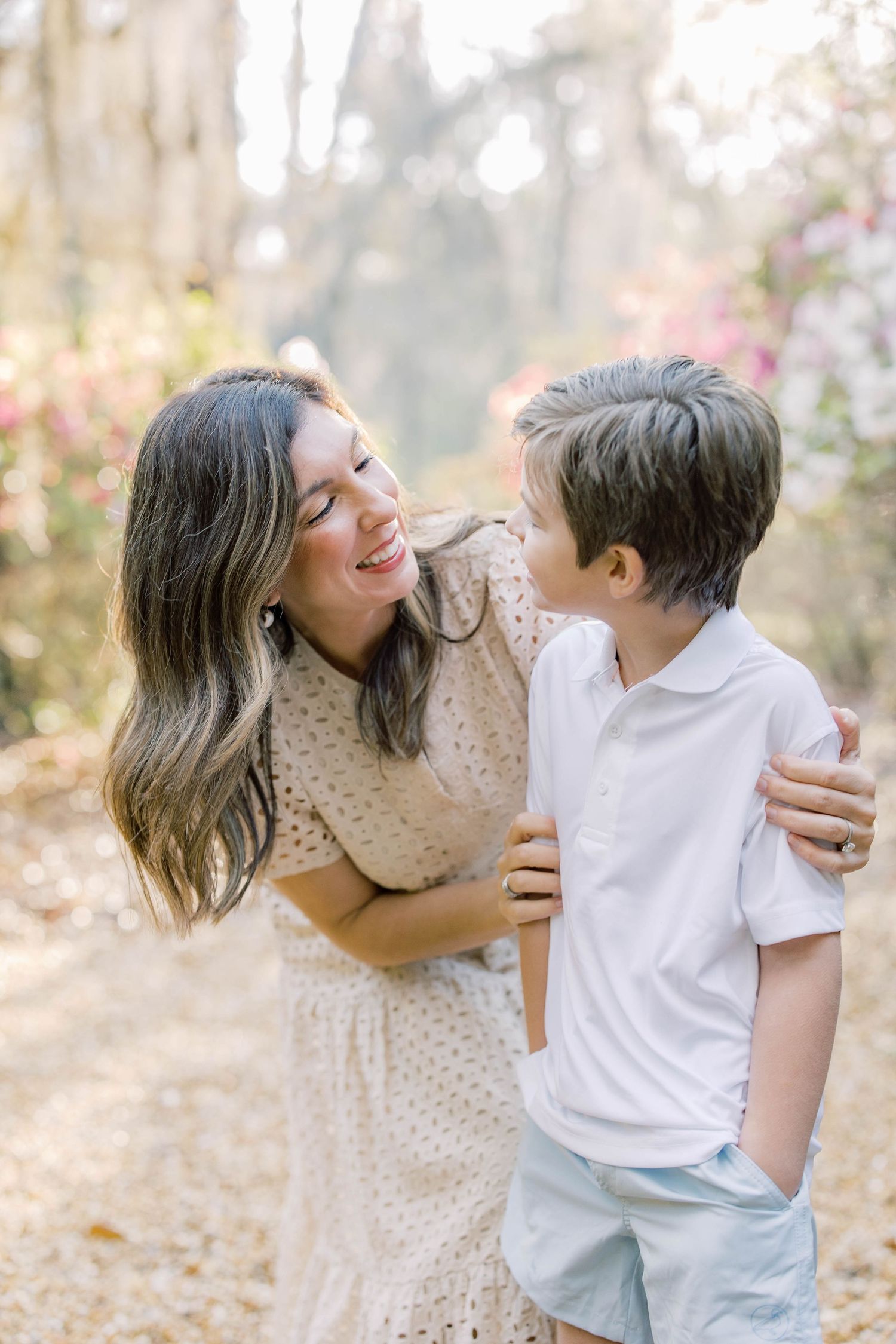 A smiling mother in a floral dress embraces her young son outdoors among blooming pink flowers.