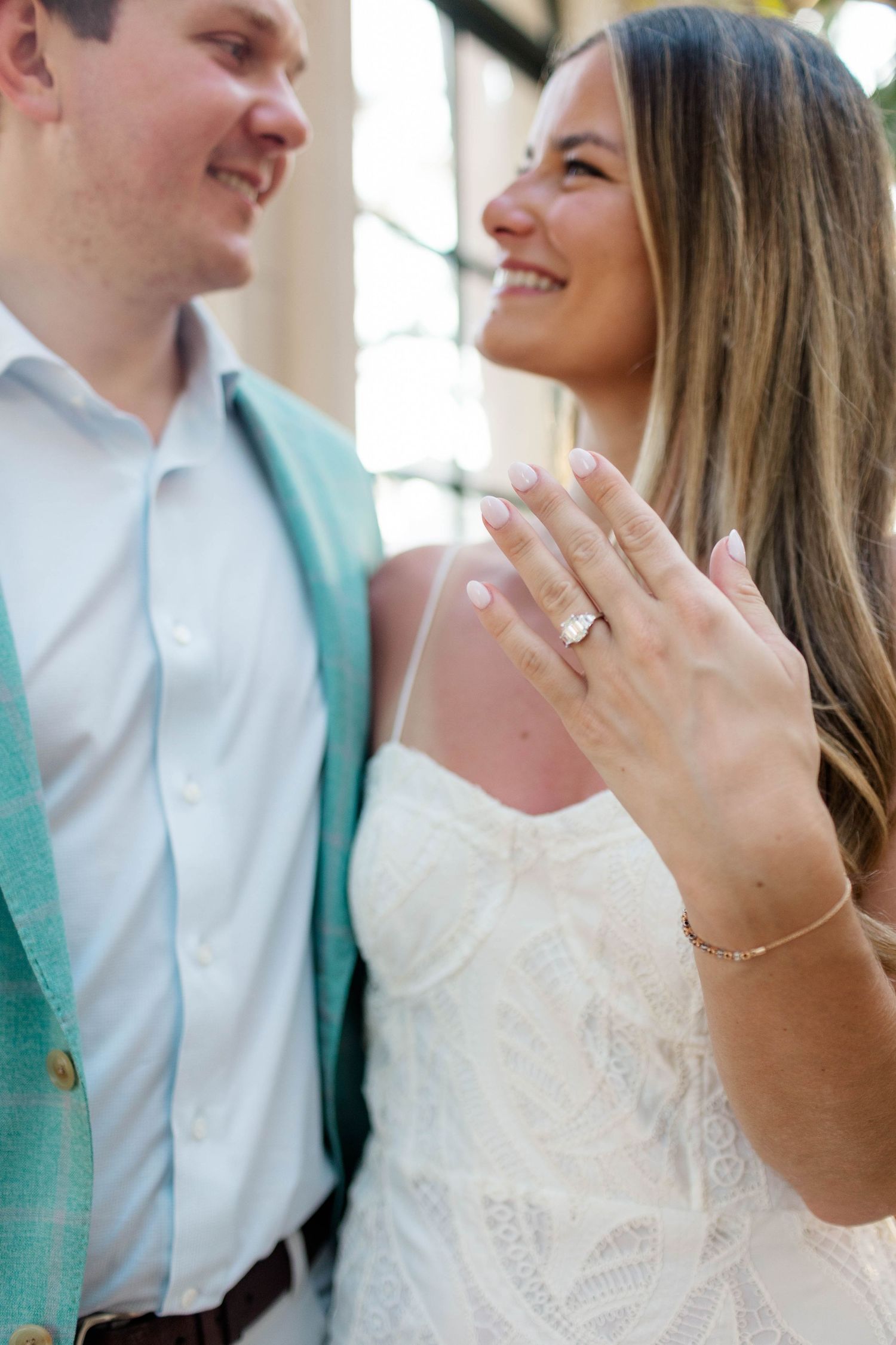 Smiling engaged couple showing off diamond engagement ring, woman in white dress with man in mint blazer.
