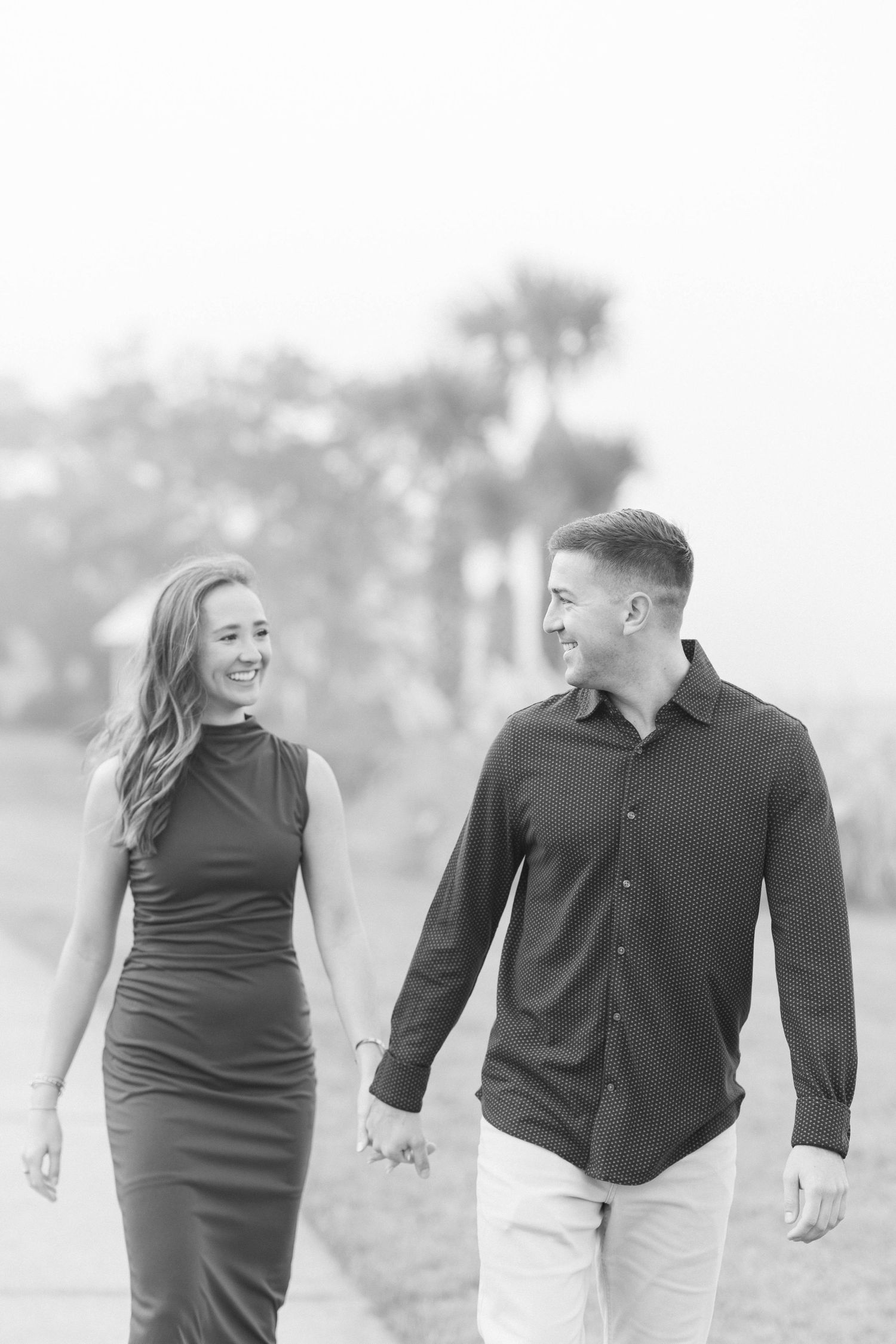 Black and white photo of a smiling couple holding hands and walking outdoors with palm trees in the background.
