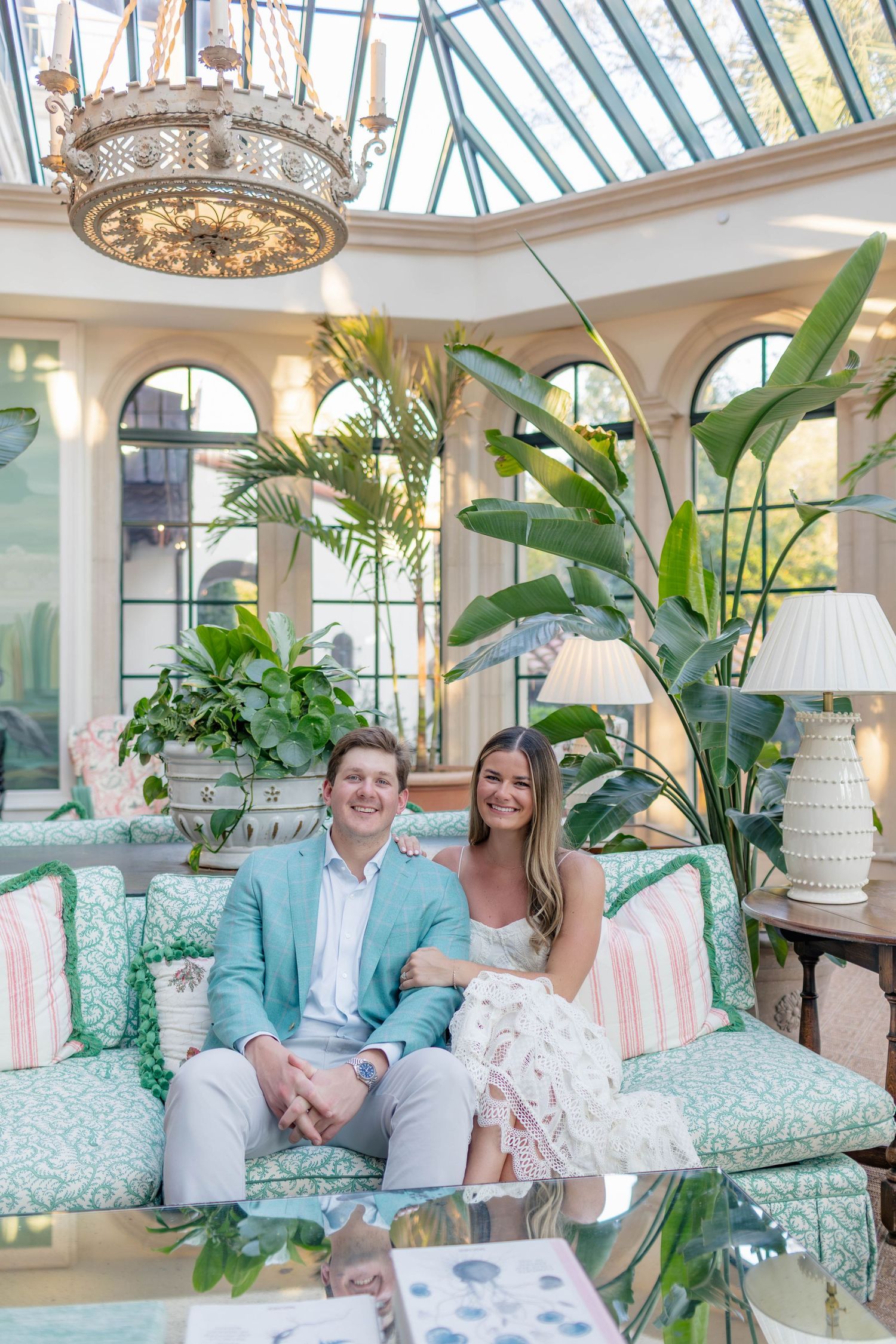 Couple sitting on a teal sofa in an elegant glass conservatory with tropical plants and a chandelier.