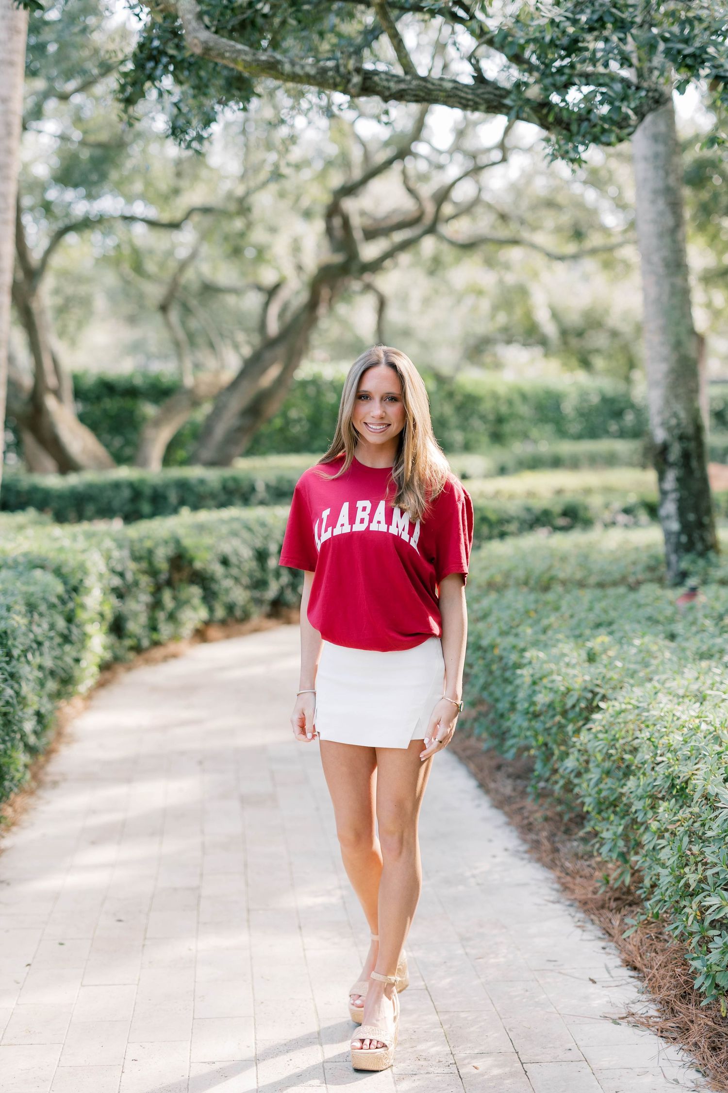 Young woman in Alabama red crop tee and white mini skirt walking on a tree-lined garden path.