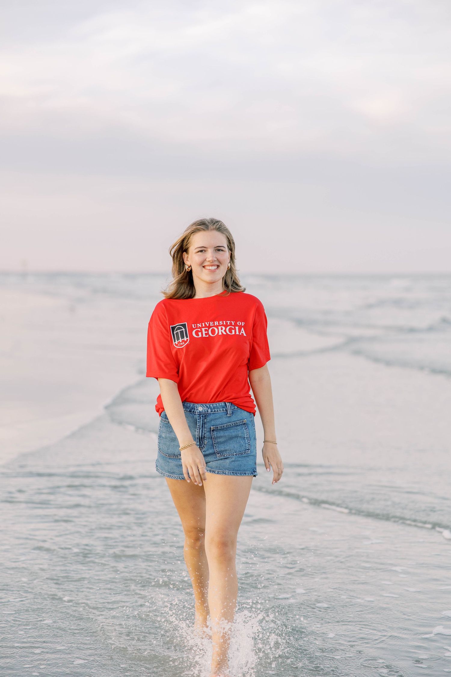 Young woman in University of Georgia red t-shirt and denim shorts smiling while wading in ocean waves at beach.