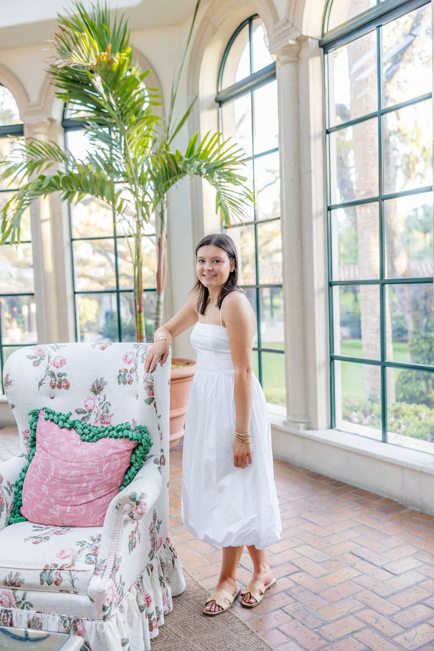 Young woman in white sundress stands beside floral armchair in sunlit conservatory with arched windows and tropical plants.