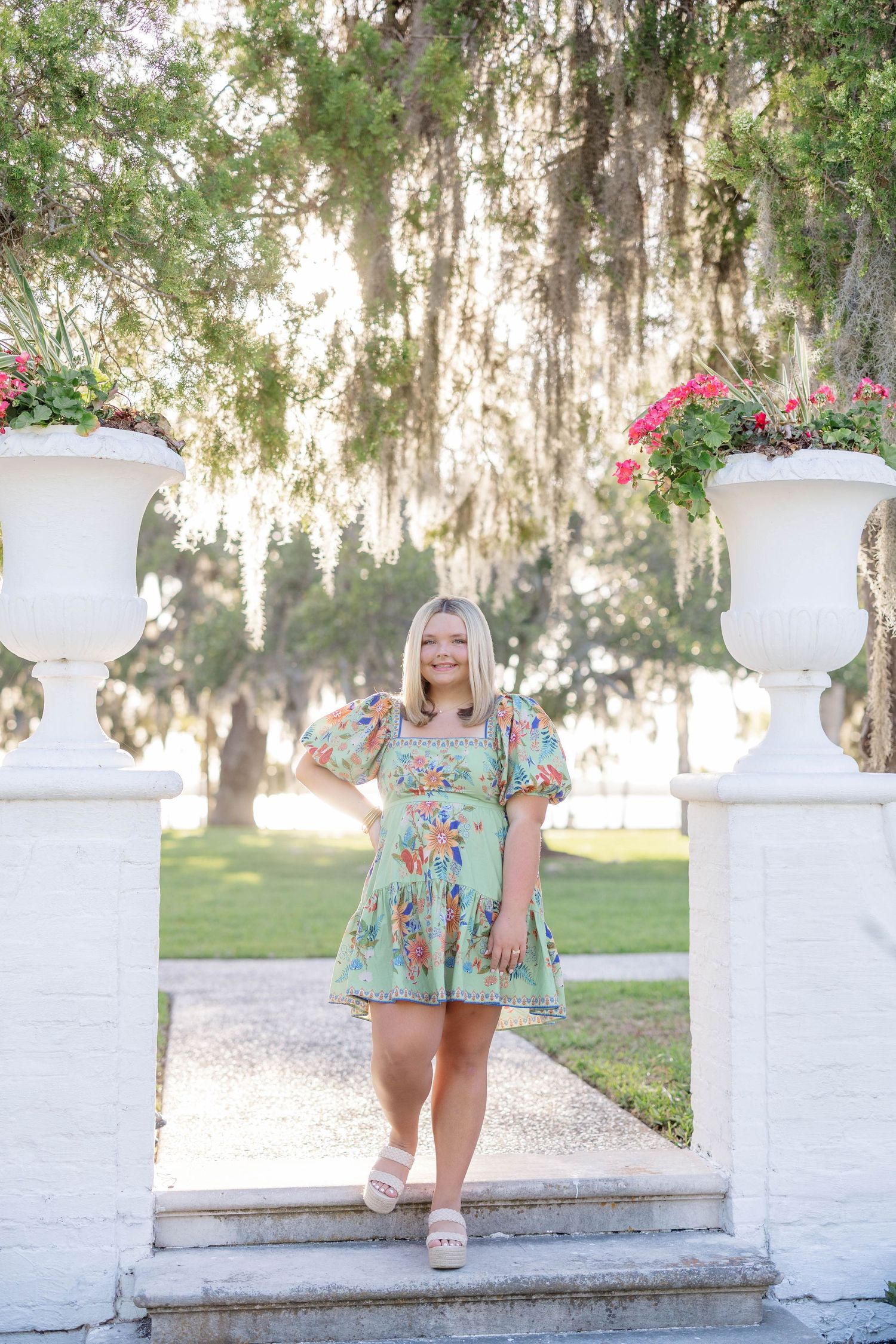 Young blonde woman in floral green dress poses between white urns with flowers, surrounded by Spanish moss-draped trees.