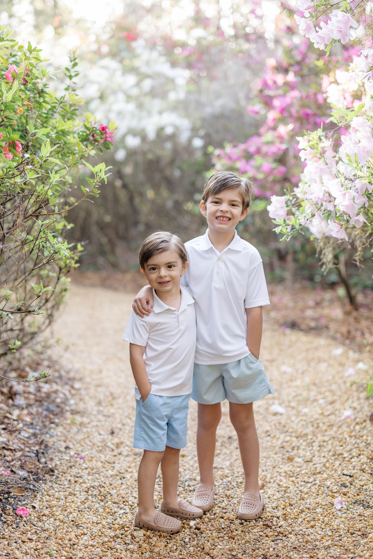 Two young brothers in matching white polos and blue shorts pose together on a gravel path among blooming flowers.