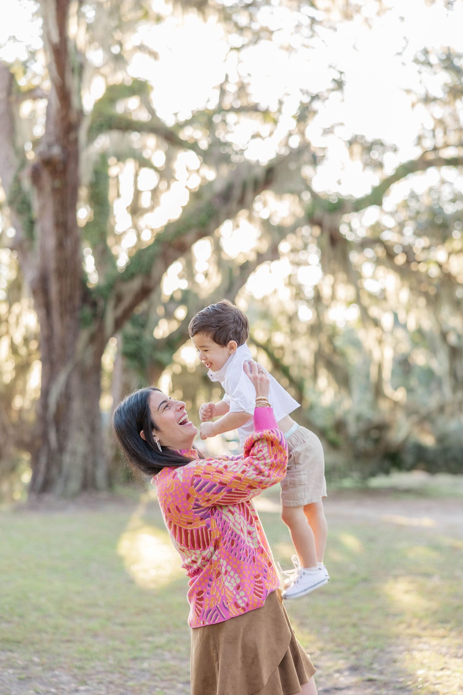 A joyful mother lifts her laughing toddler outdoors under moss-draped oak trees, both smiling brightly.