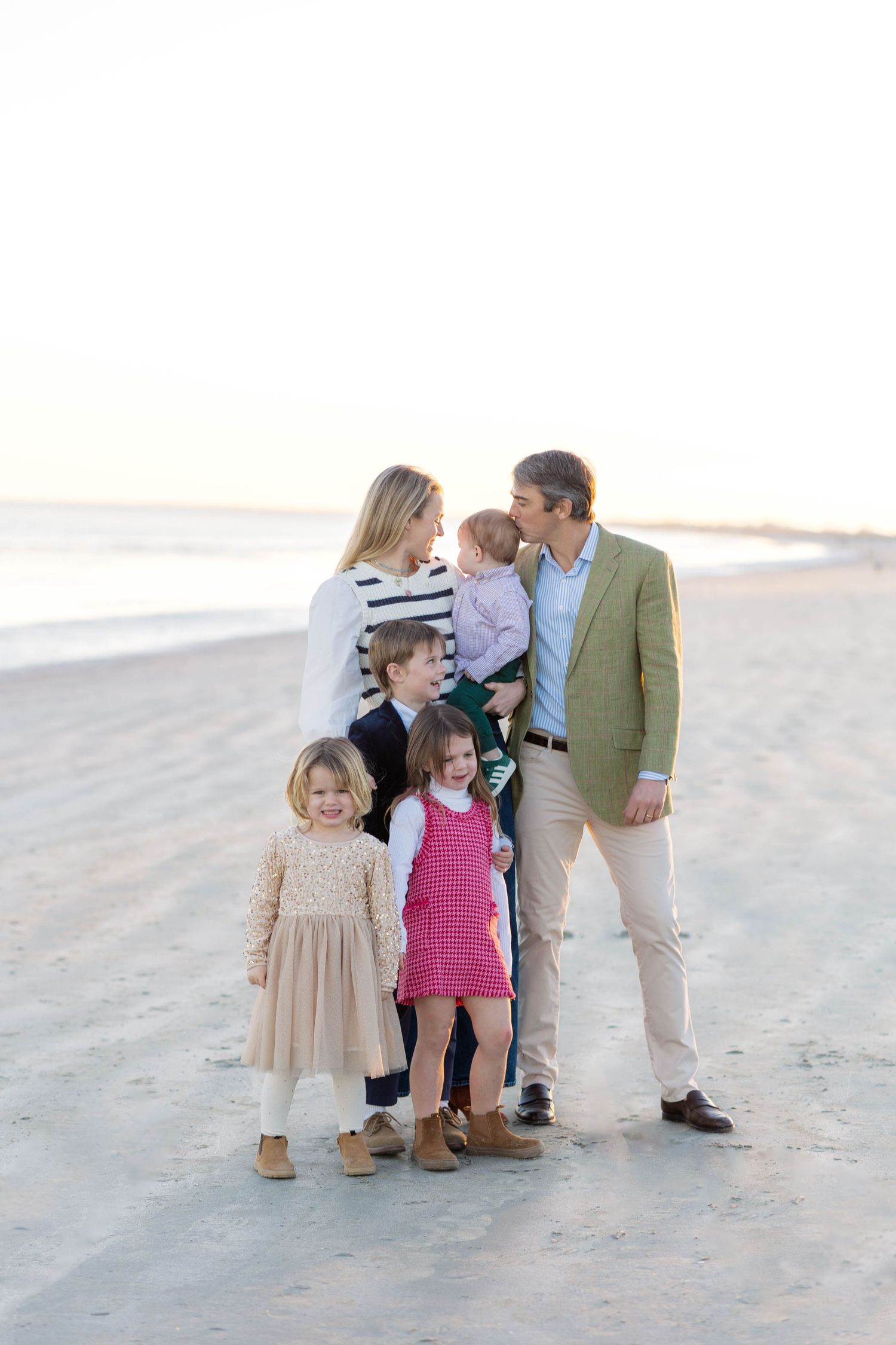 Family of six posing on a sandy beach at sunset, parents holding baby while three young children stand in front.