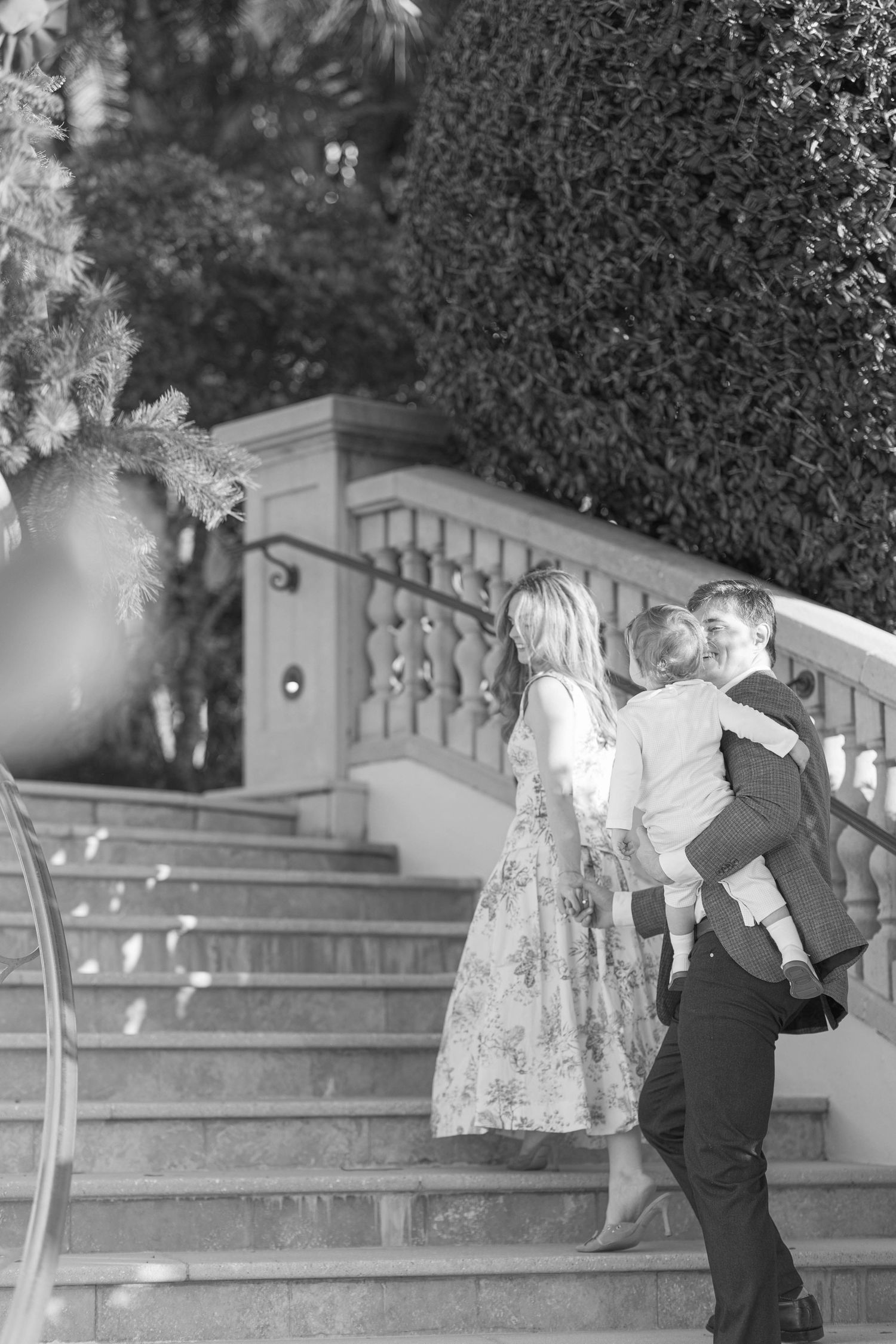 Black and white photo of a family walking up elegant stone stairs, man carrying child while woman in floral dress looks on.