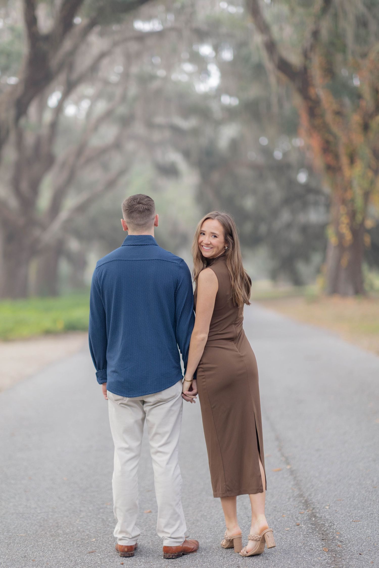 Couple holding hands on a tree-lined foggy road, woman in brown dress smiling over shoulder.