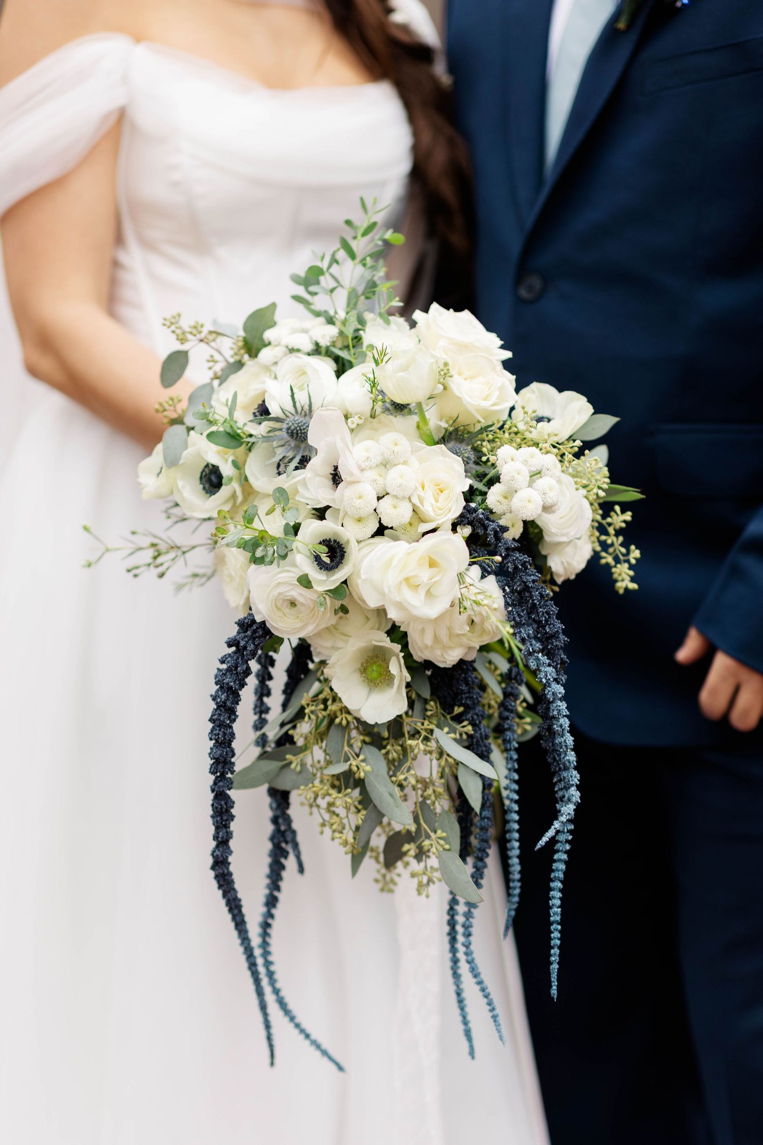 A stunning white bridal bouquet features elegant white roses and delicate blue delphiniums against a navy and white background.