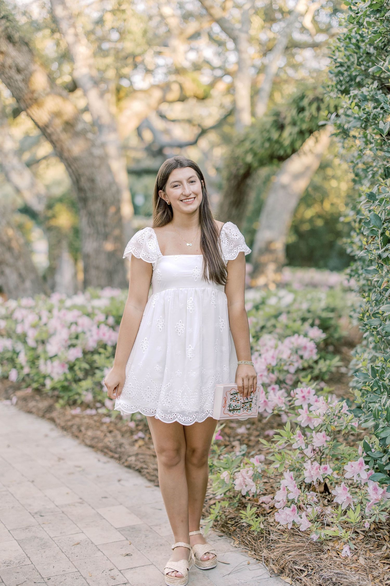 Young woman in white eyelet dress walking along a garden path with pink flowering bushes.