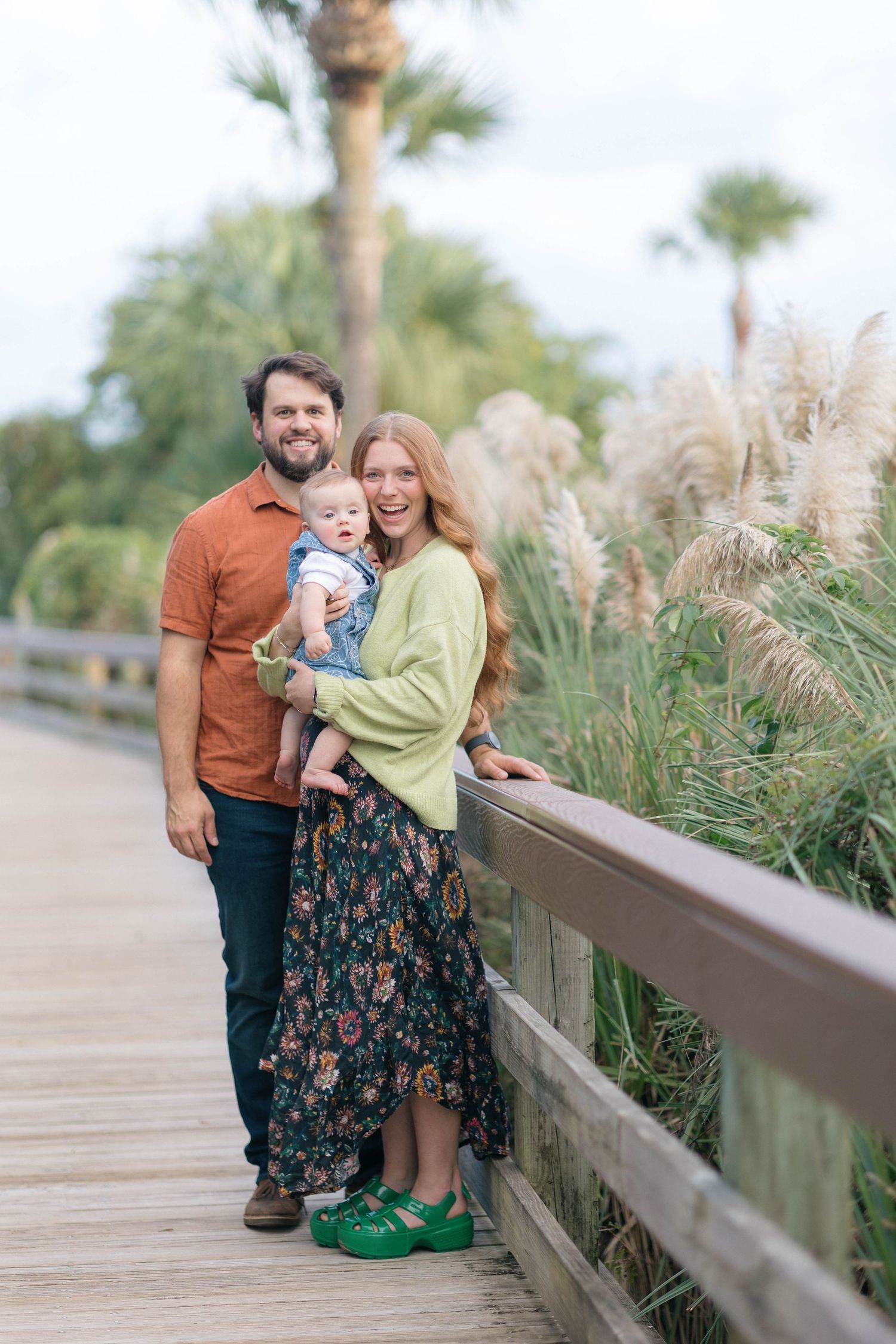 A smiling family of three poses on a wooden boardwalk surrounded by lush greenery and palm trees.