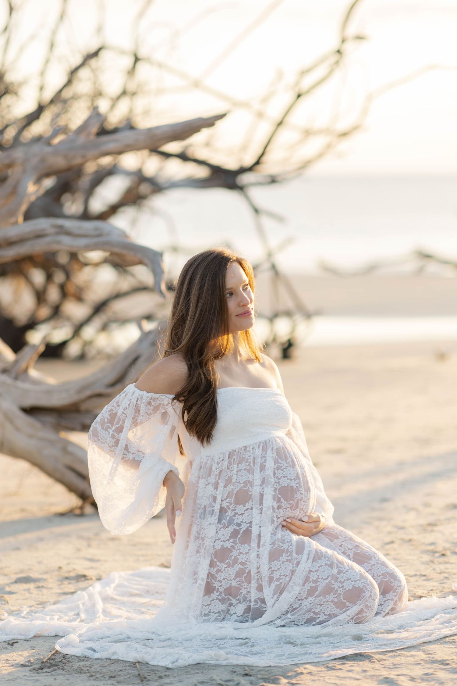 Pregnant woman in white lace off-shoulder gown kneels on sandy beach near driftwood at golden hour.