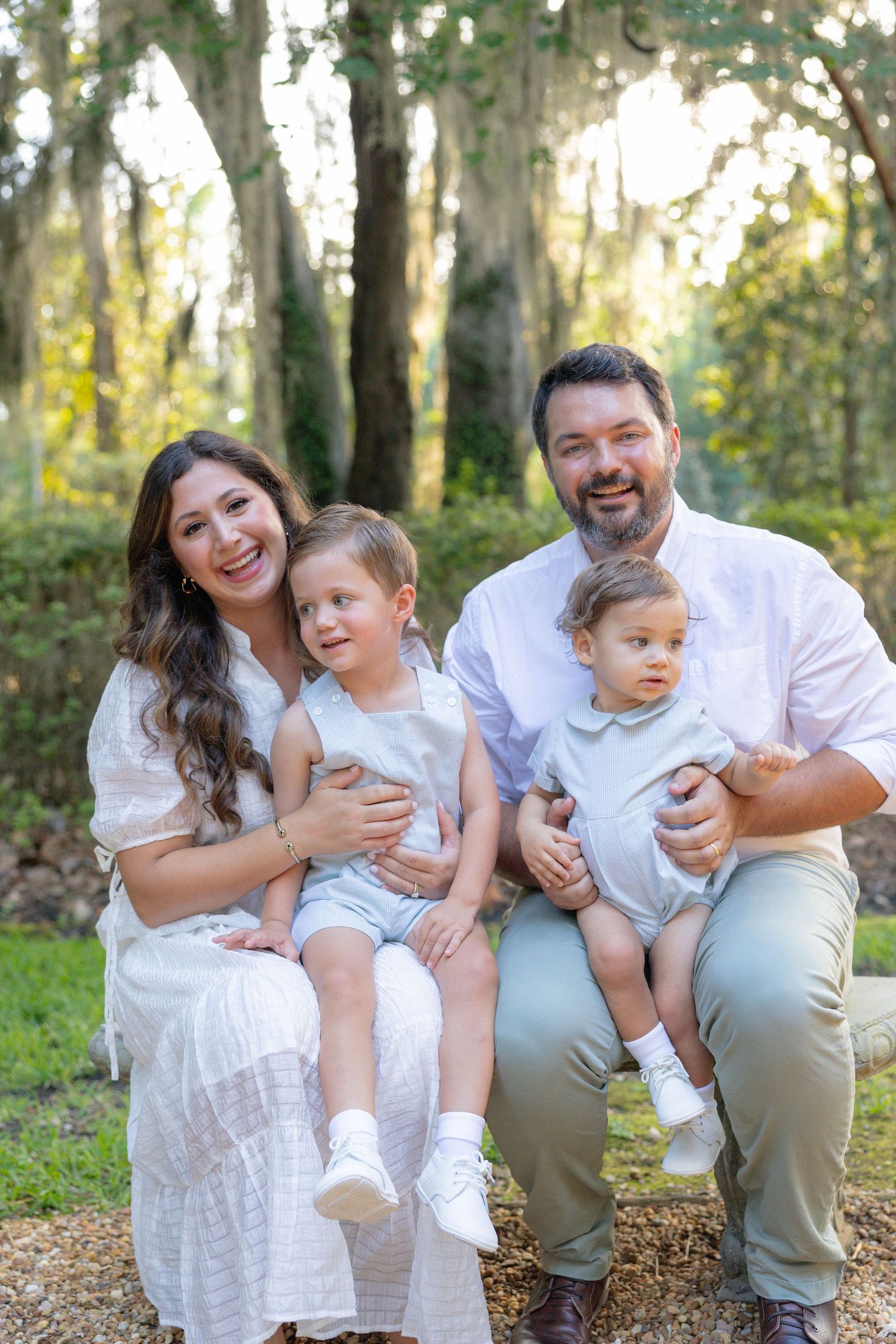 Happy family of four posing outdoors among trees, parents holding two young children dressed in white and blue.