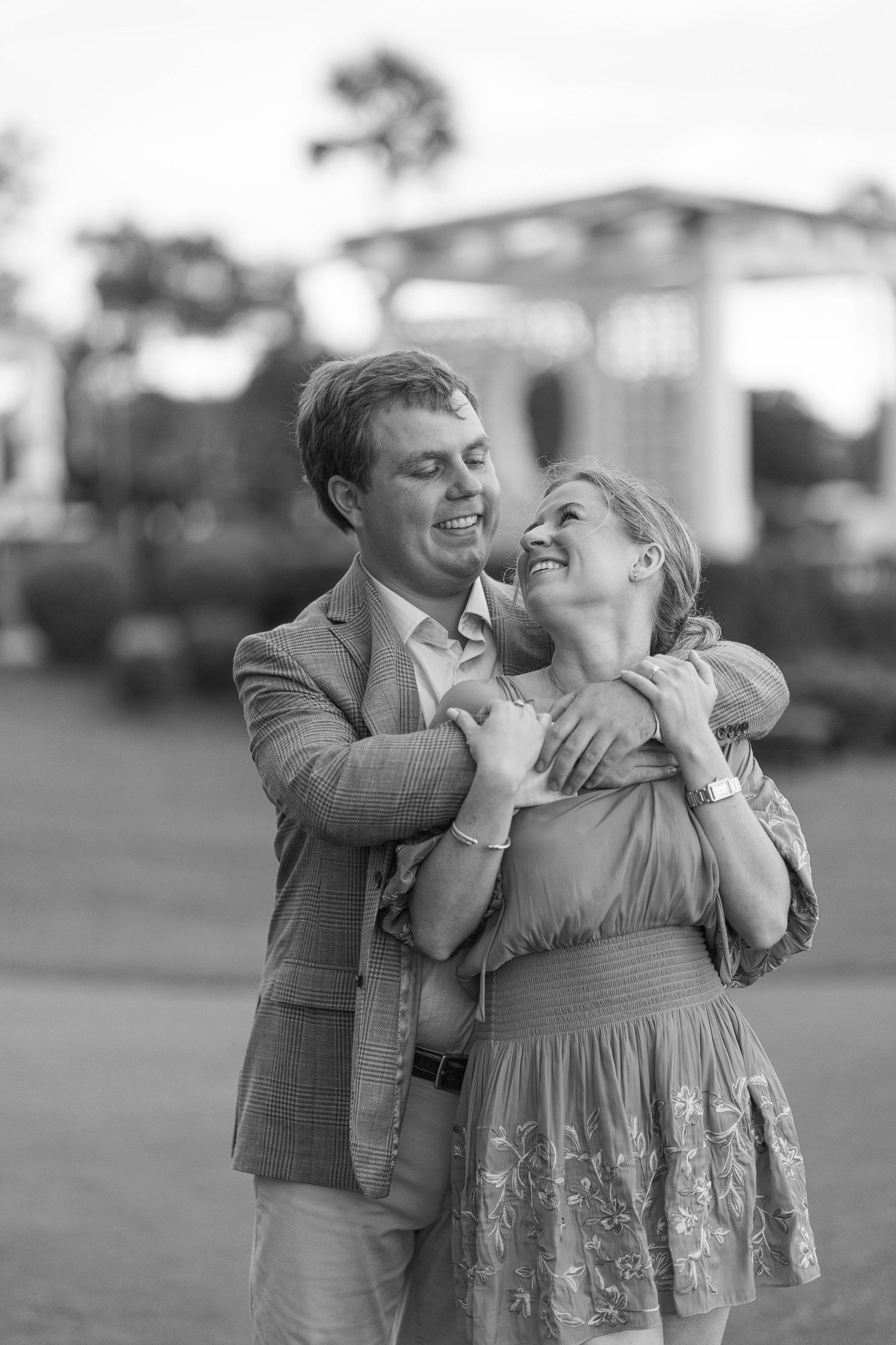 Black and white photo of a happy couple embracing outdoors, smiling at each other in a park setting.