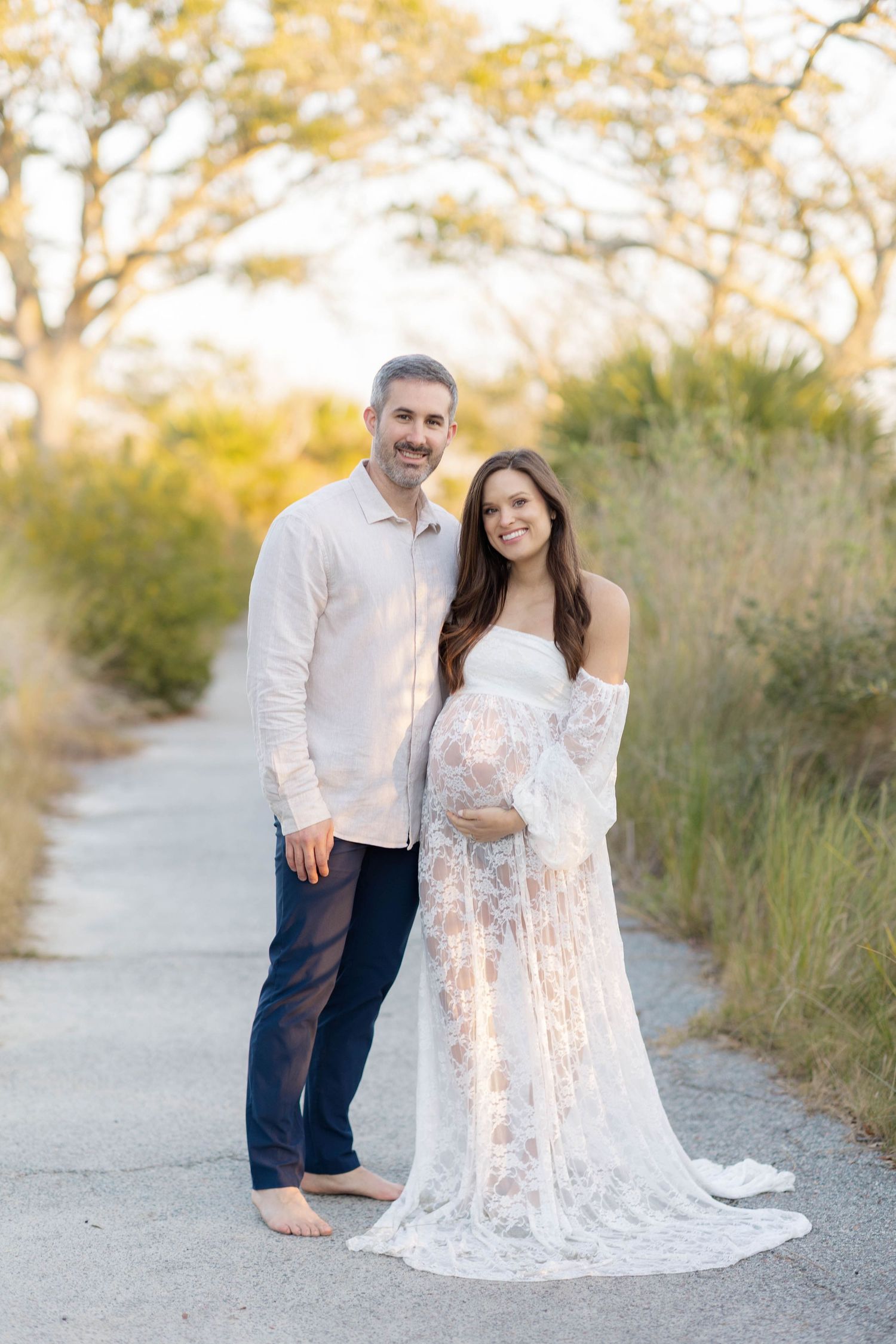 Couple poses outdoors for maternity photo, woman in white lace gown cradling baby bump, man in white shirt and navy pants.