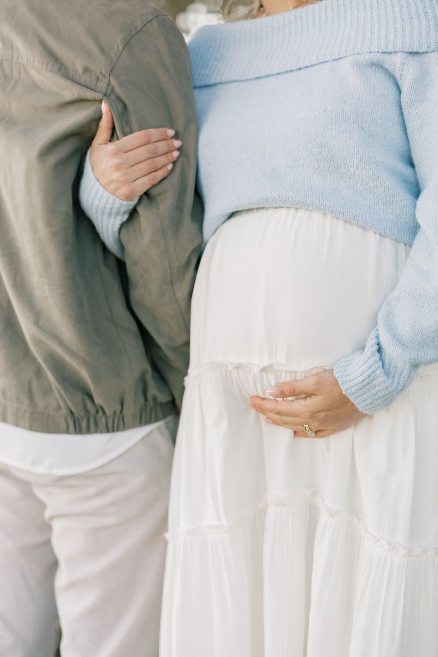 Pregnant woman in blue off-shoulder sweater and white skirt stands beside partner in olive jacket during maternity photoshoot.