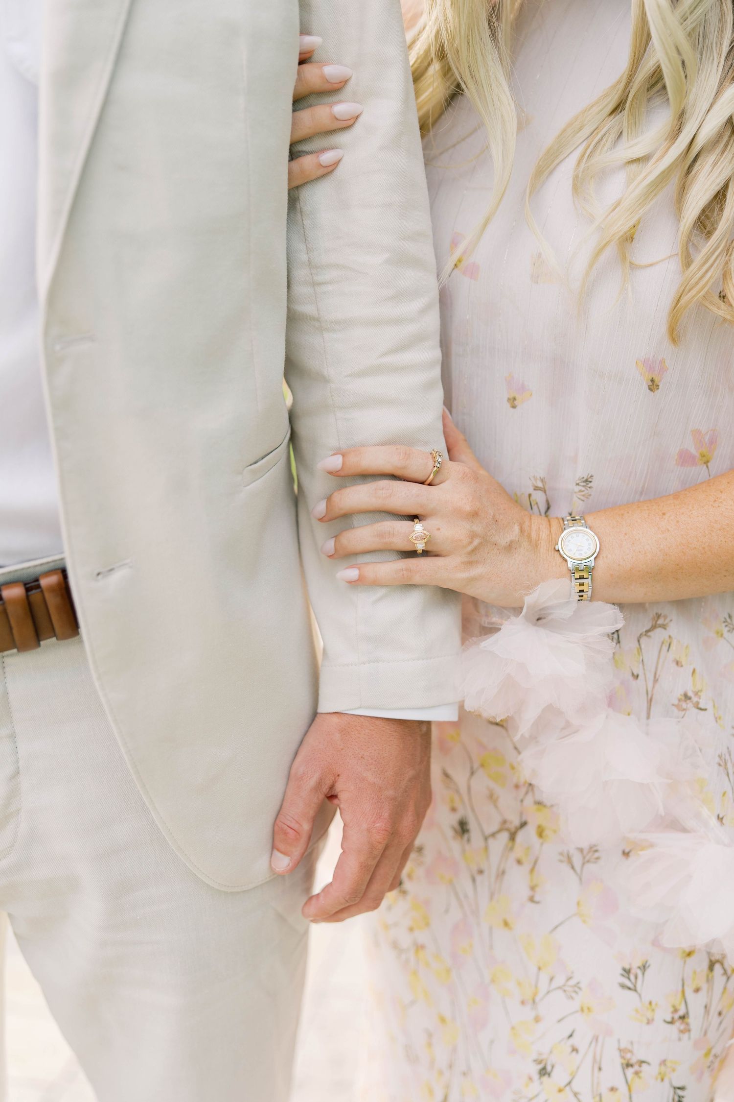 Couple posing closely, woman in floral dress with ring and gold watch holding man's arm in light gray suit.