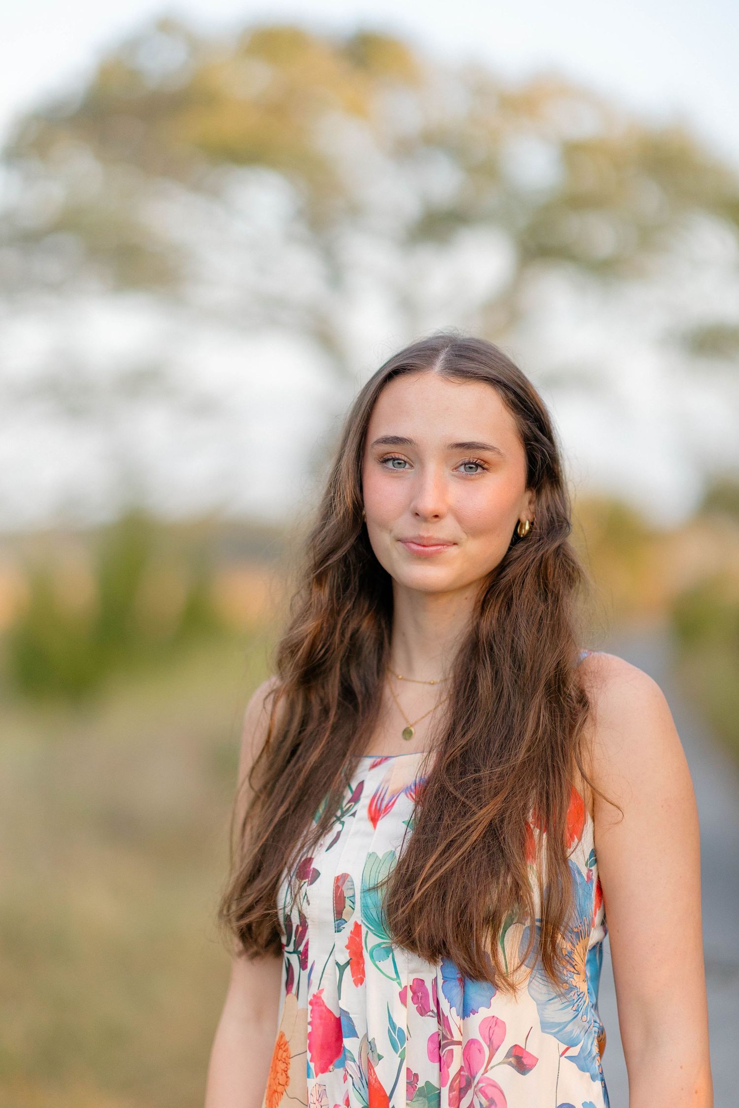 Young woman with long brown hair wearing a colorful floral dress outdoors in natural light.