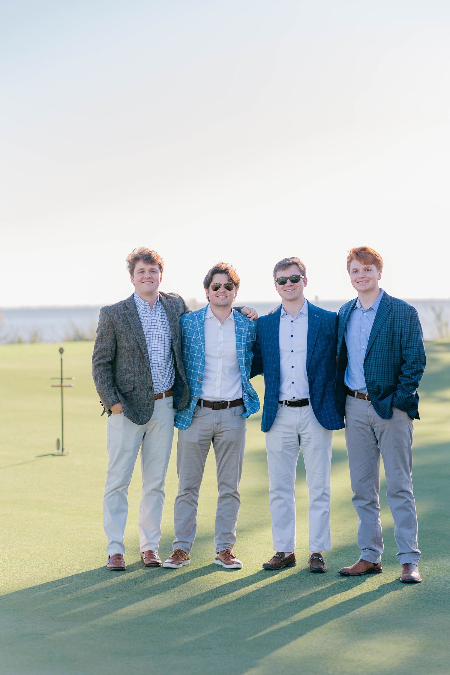 Four young men in smart casual blazers posing together on a golf course green.
