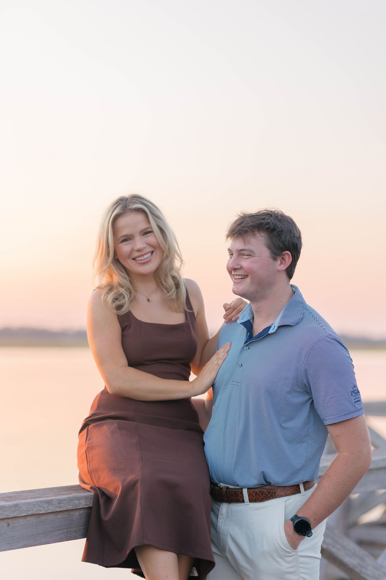 A smiling couple poses on a waterfront dock at sunset, woman in brown dress with arm around man in blue polo shirt.