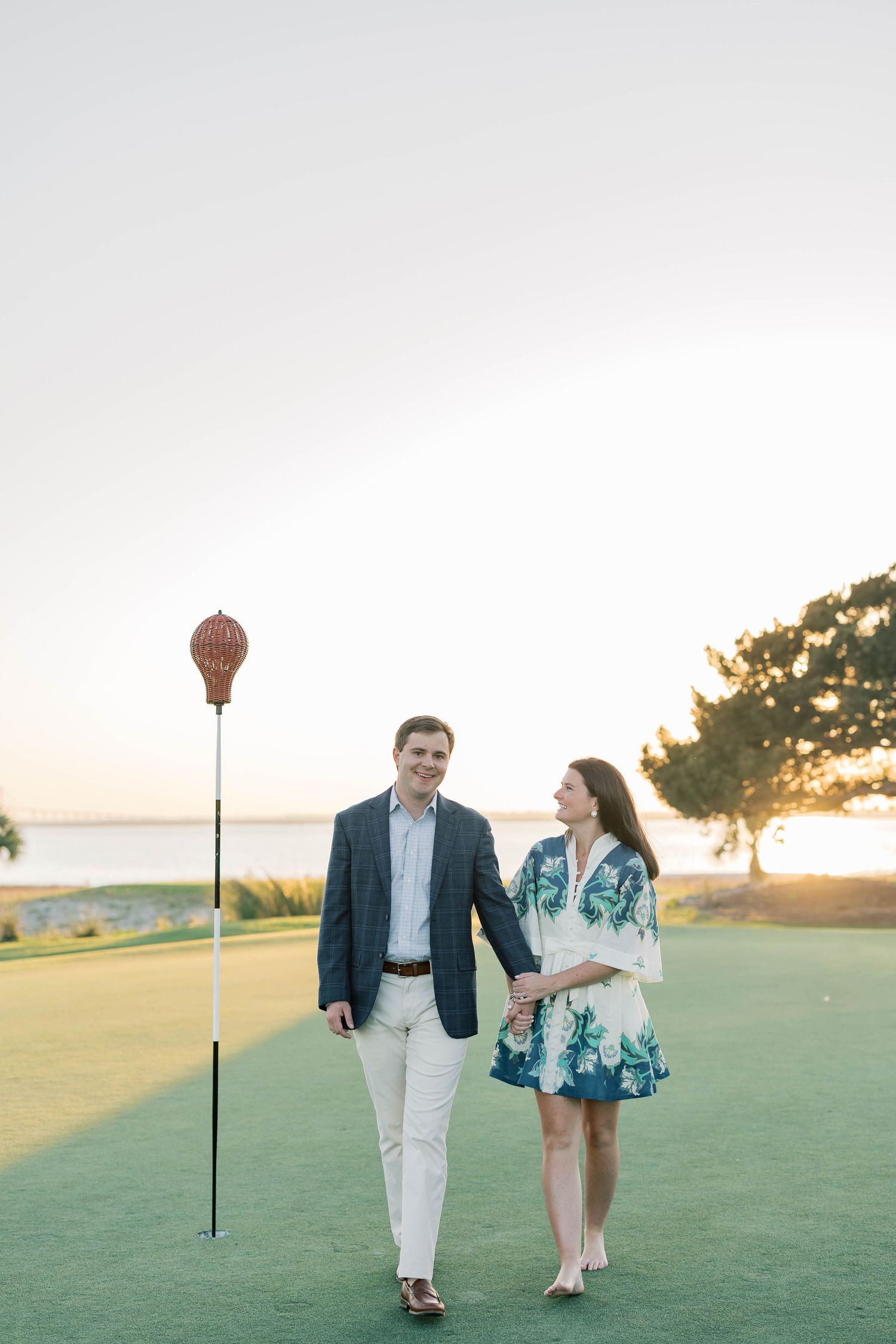 Couple holding hands walking on a golf course green at sunset, man in blazer and woman in floral dress.
