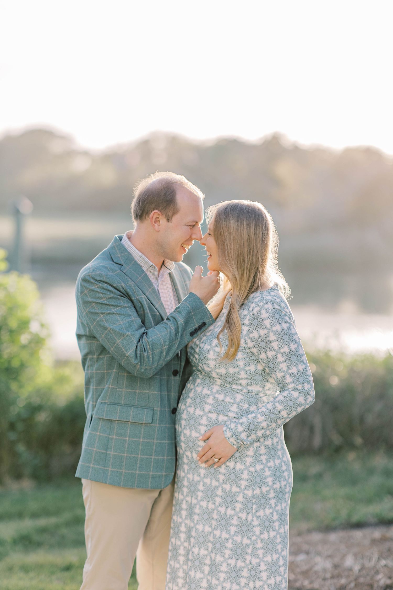 A couple shares a tender moment outdoors at golden hour, the woman in a floral dress cradling her baby bump.