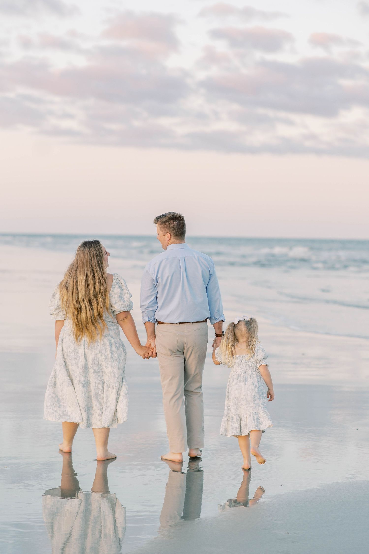 Family of three walking hand-in-hand on a serene beach at sunset, reflections visible on wet sand.