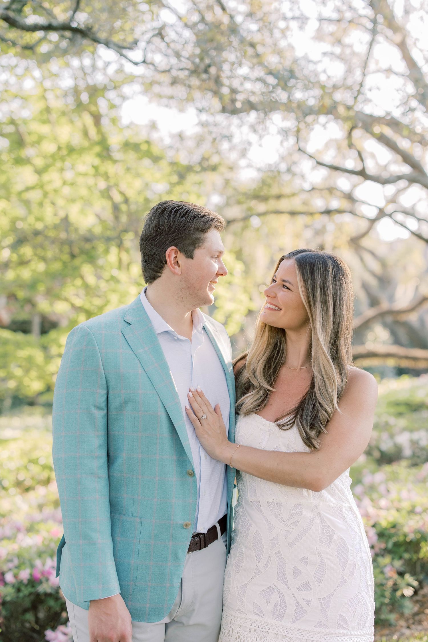 A smiling couple poses outdoors among blooming trees, the woman in white lace and man in a light blue blazer.