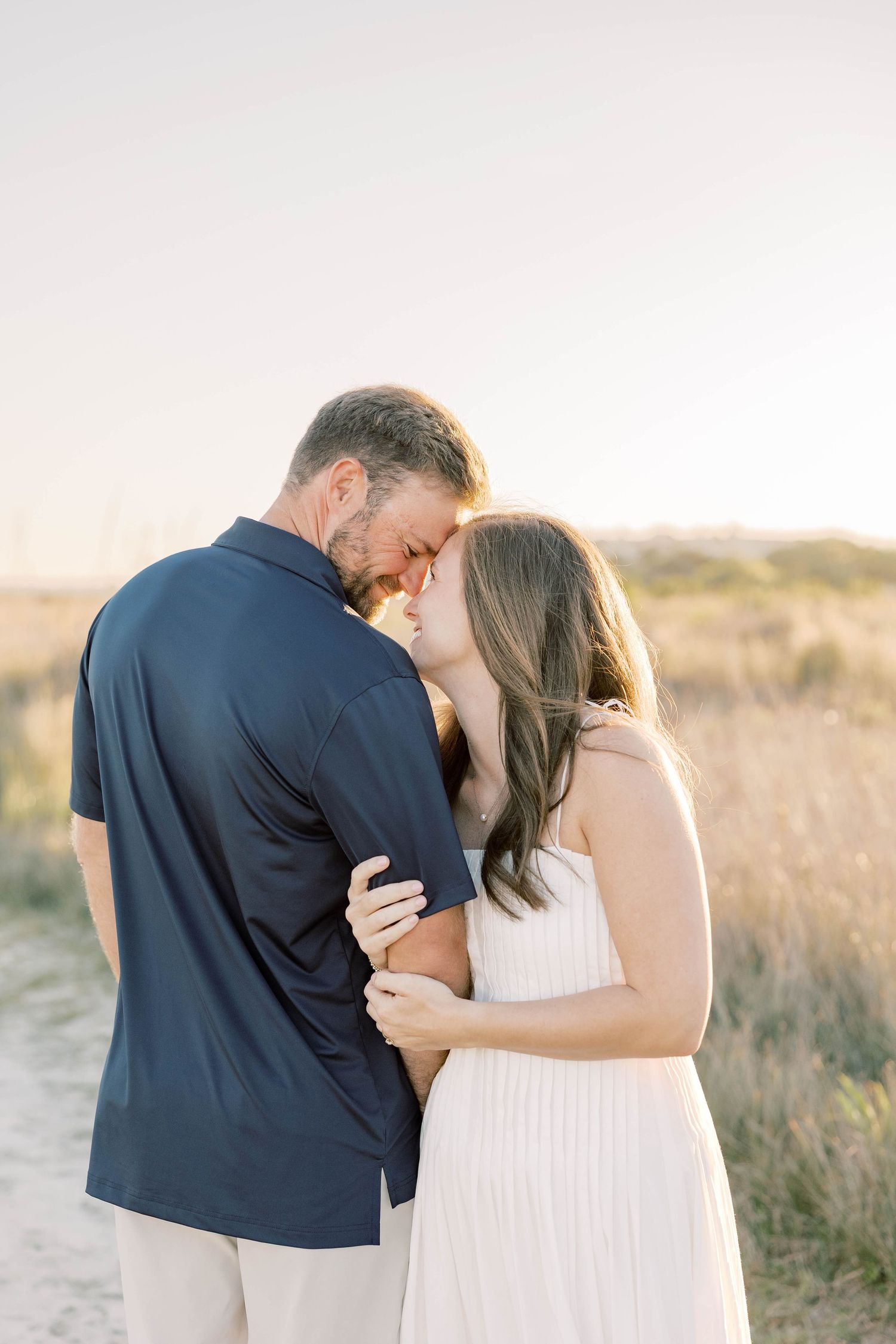 Couple sharing a romantic forehead touch on a sandy beach path at golden hour, man in navy polo, woman in white dress.