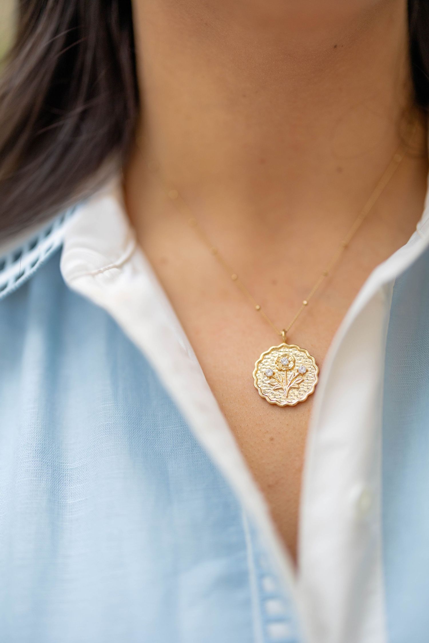 Close-up of gold floral coin pendant necklace on woman wearing light blue button-down shirt.