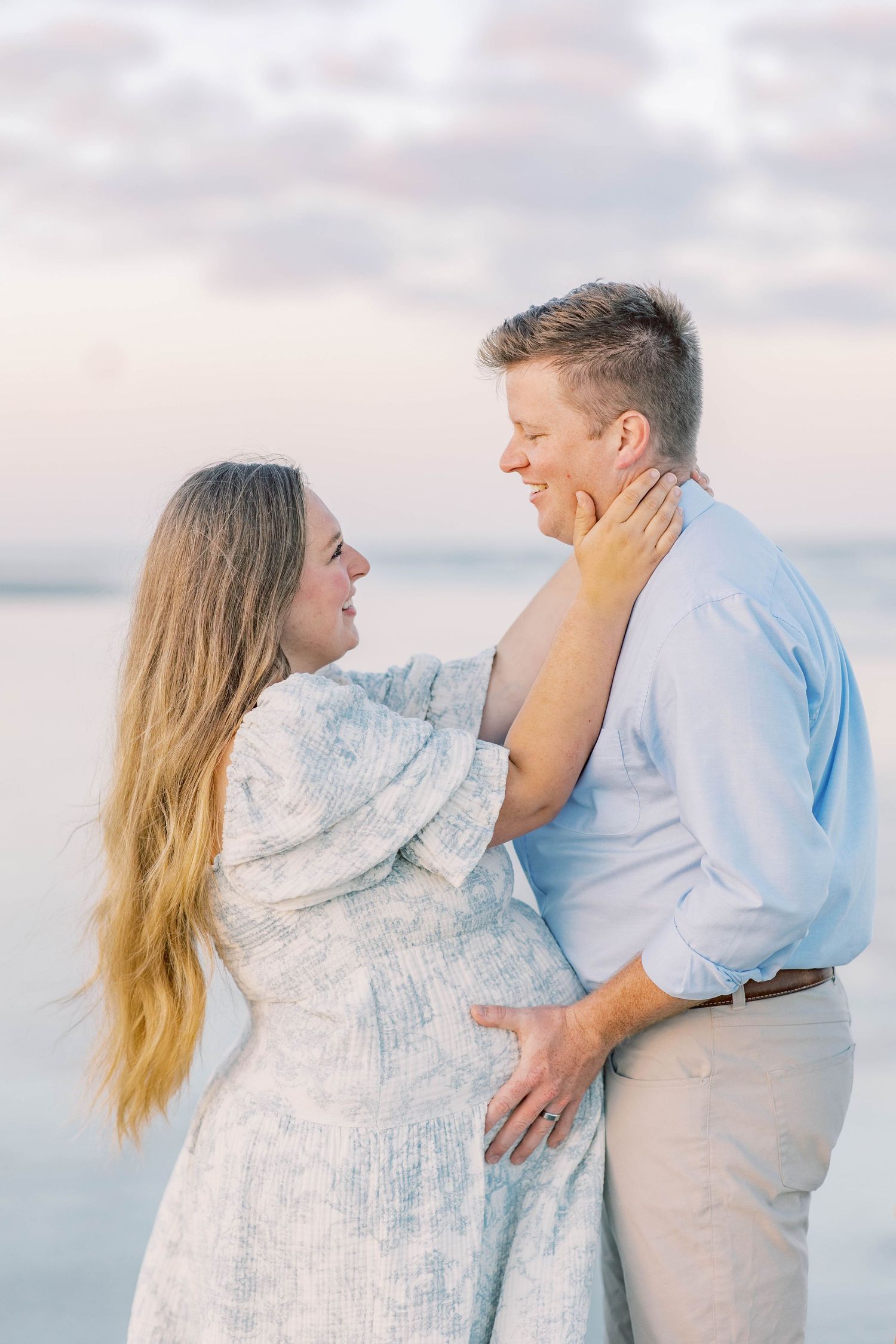 Pregnant couple embracing on beach at sunset, woman in floral dress touching man's face, soft pastel sky background.
