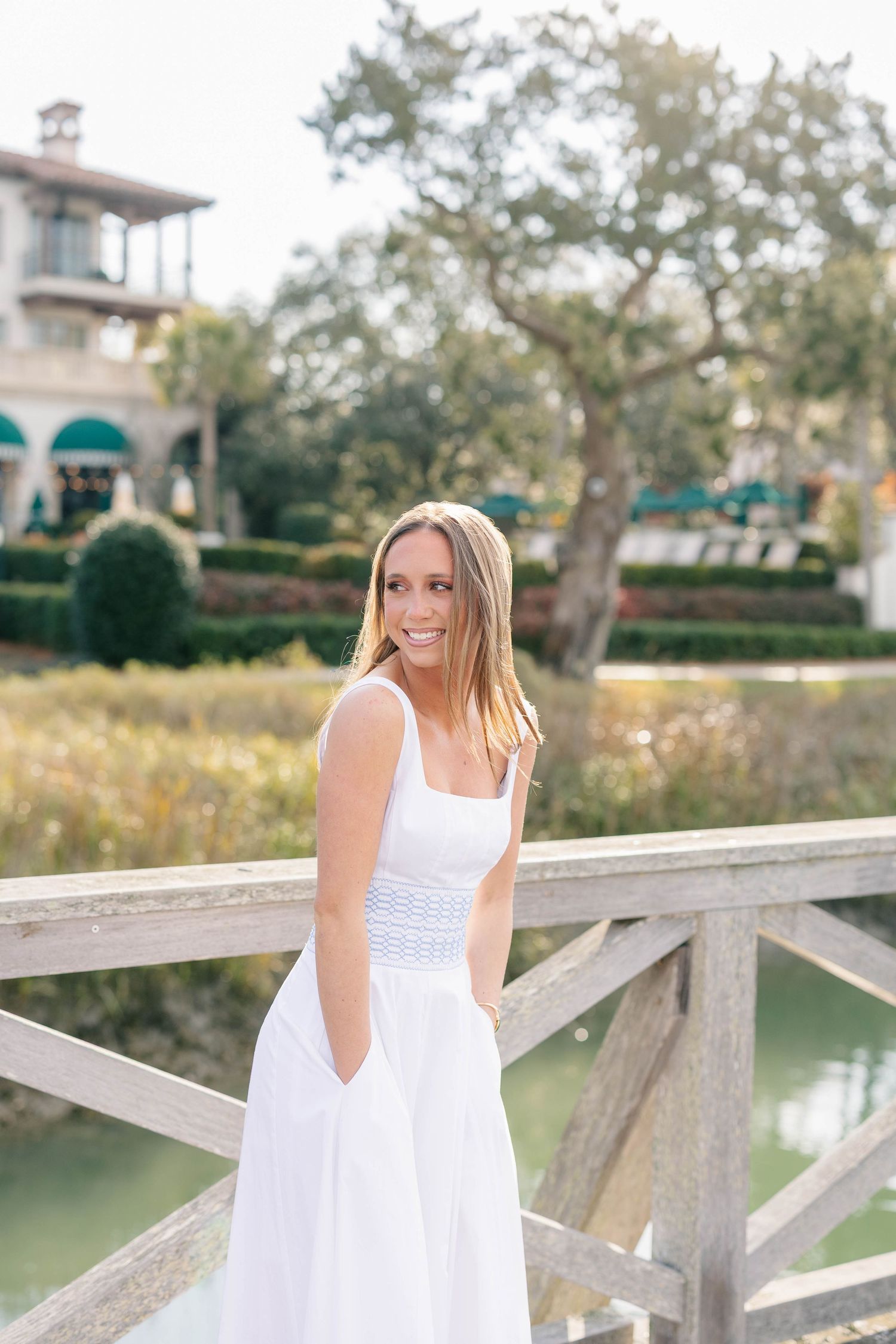 Woman in white dress posing by wooden railing near waterfront with buildings in background.