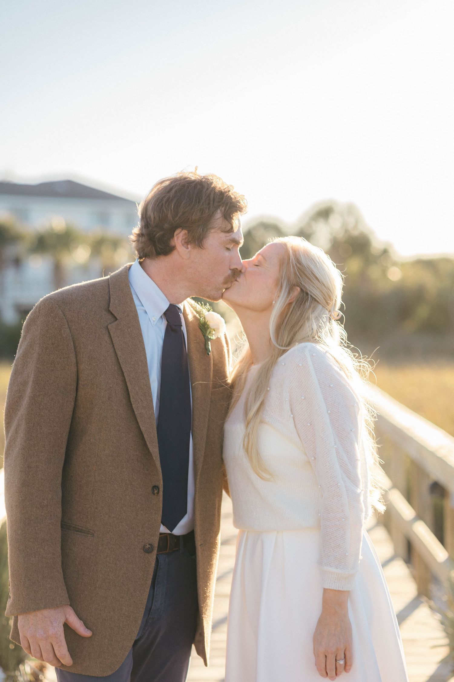 A romantic couple shares a kiss in a golden sunset setting with a brown tweed blazer and white dress.