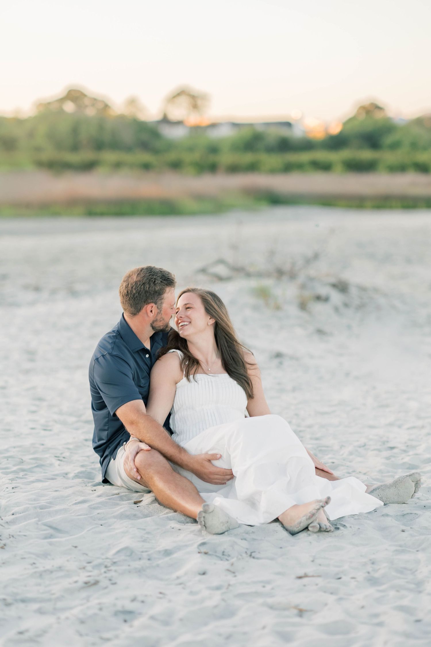 A couple sits together on sandy beach, woman in white dress smiling as man whispers to her at golden hour.