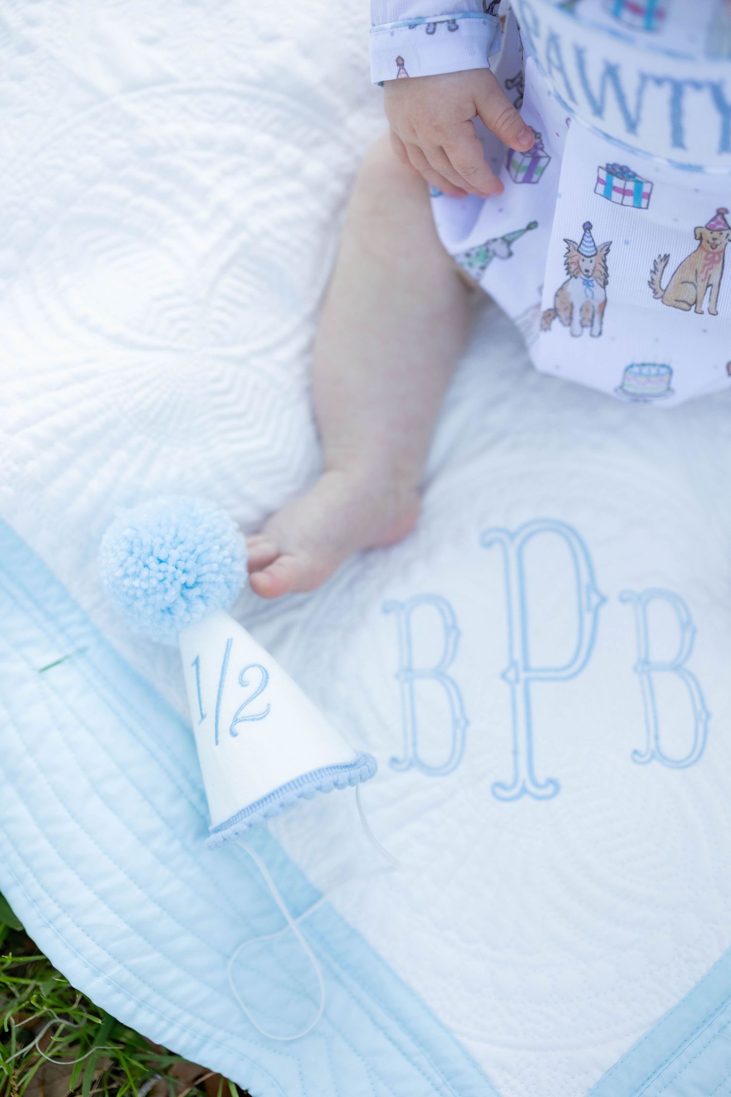 Baby's bare feet beside a half birthday party hat on a monogrammed BPB white quilted blanket.