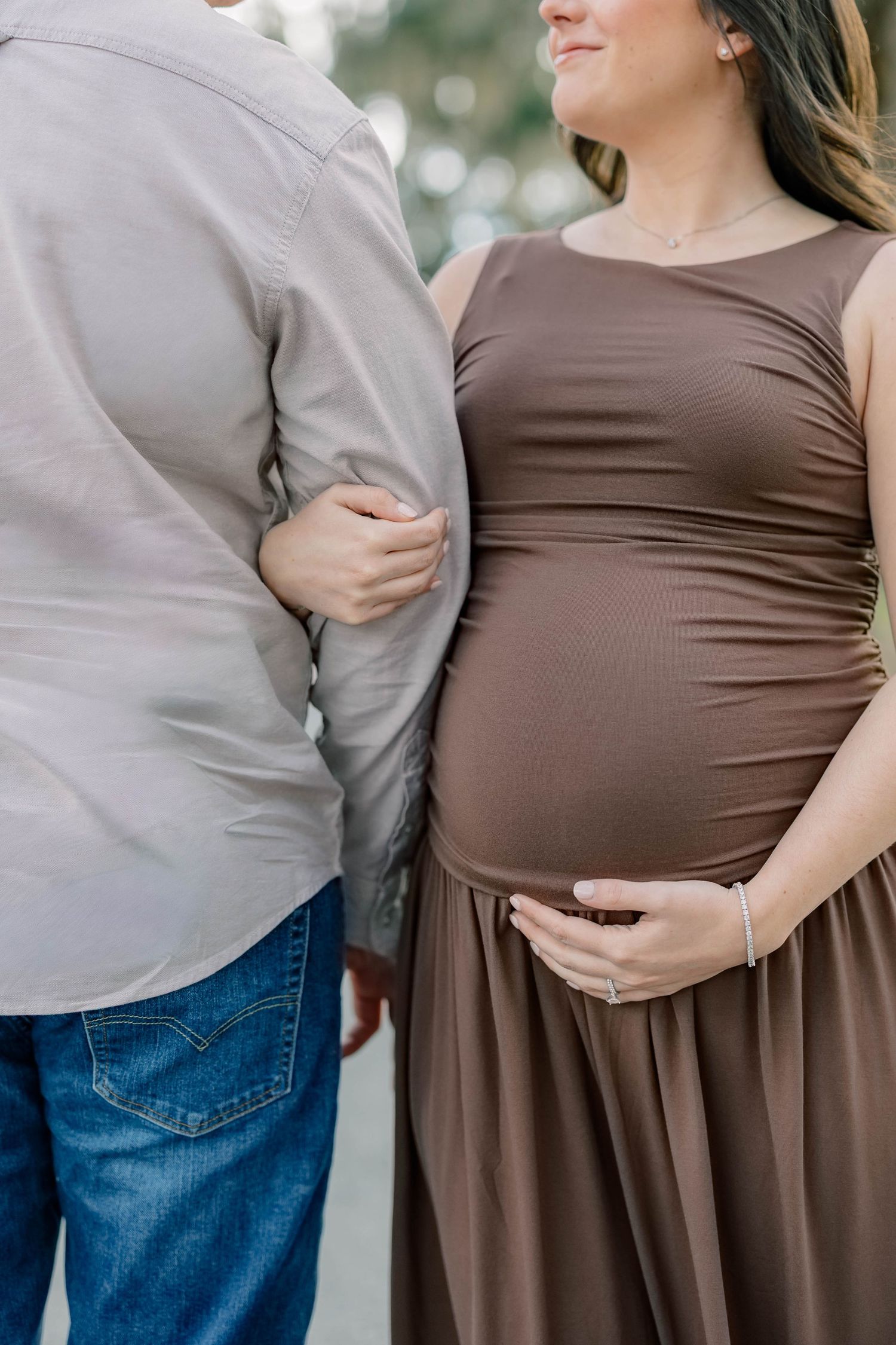Expectant couple embraces tenderly outdoors, wearing brown dress and grey shirt, with hands resting on pregnant belly.