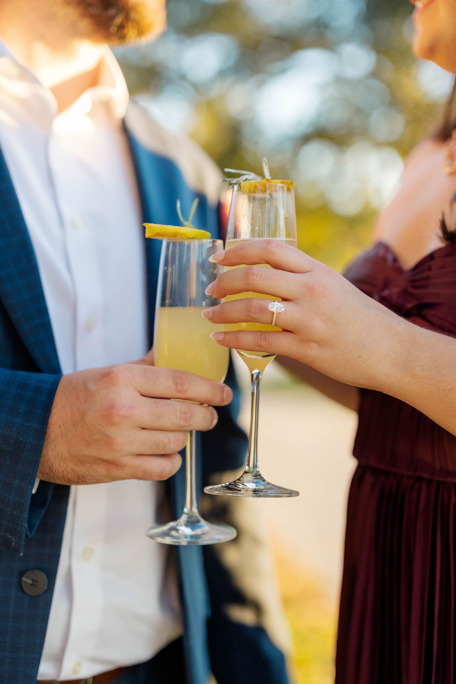 Couple toasting with champagne flutes garnished with lemon, woman wearing diamond engagement ring, outdoor celebration.