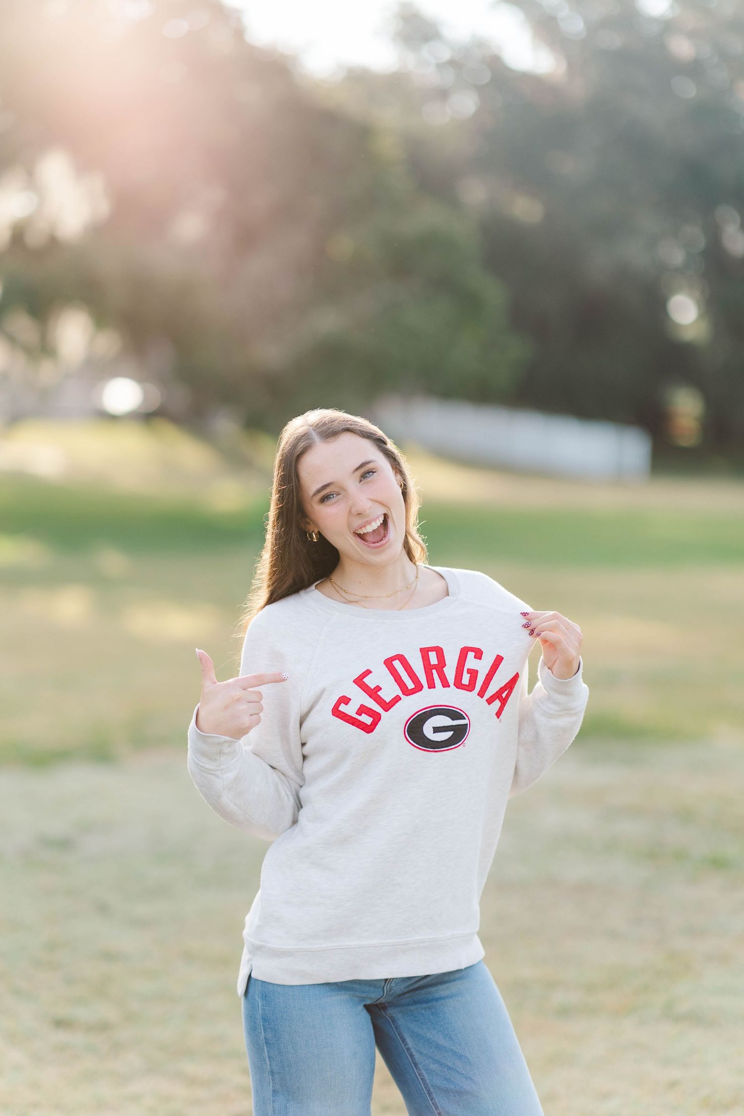 Young woman proudly wearing a Georgia Bulldogs sweatshirt, pointing at logo, smiling outdoors in warm sunlight.