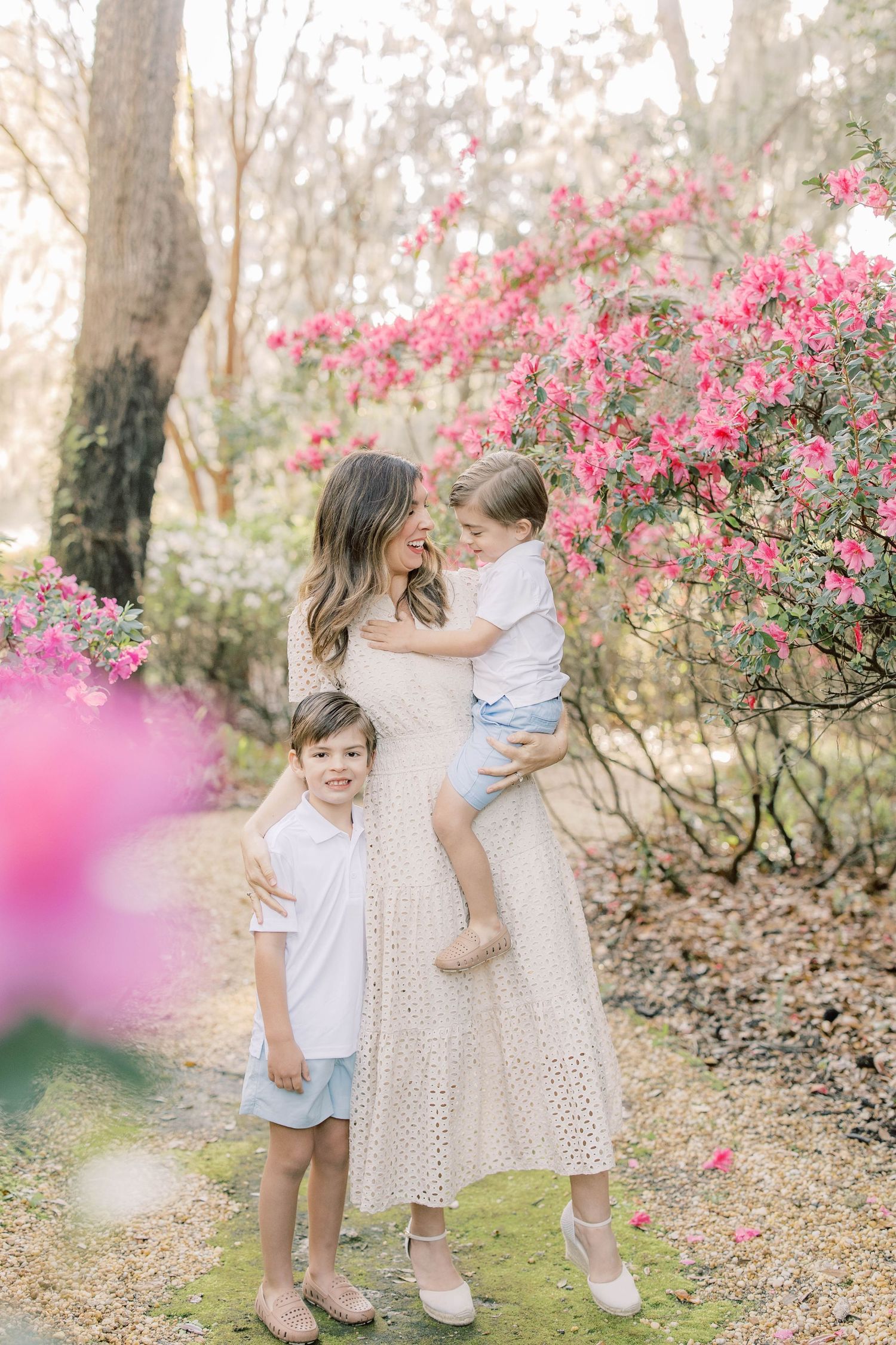 Mother in white dress holds toddler while young boy stands beside her among pink azalea blooms in spring garden.