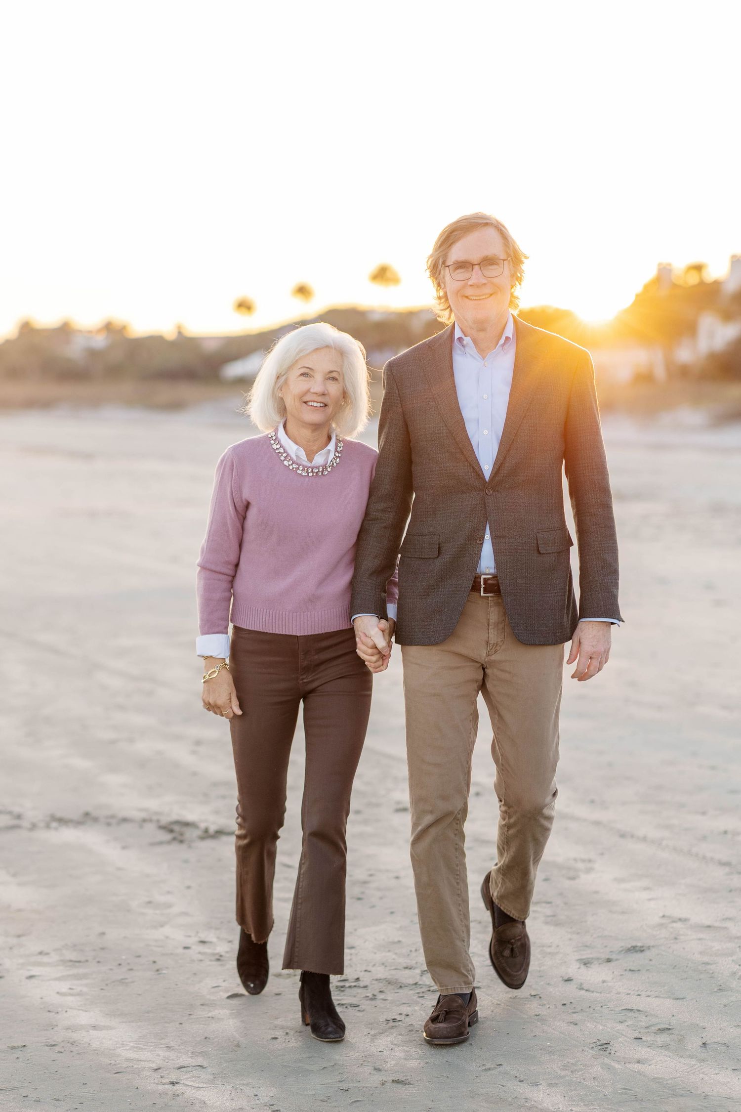 Elderly couple holding hands walking on a sandy beach at sunset, woman in pink sweater and man in blazer.