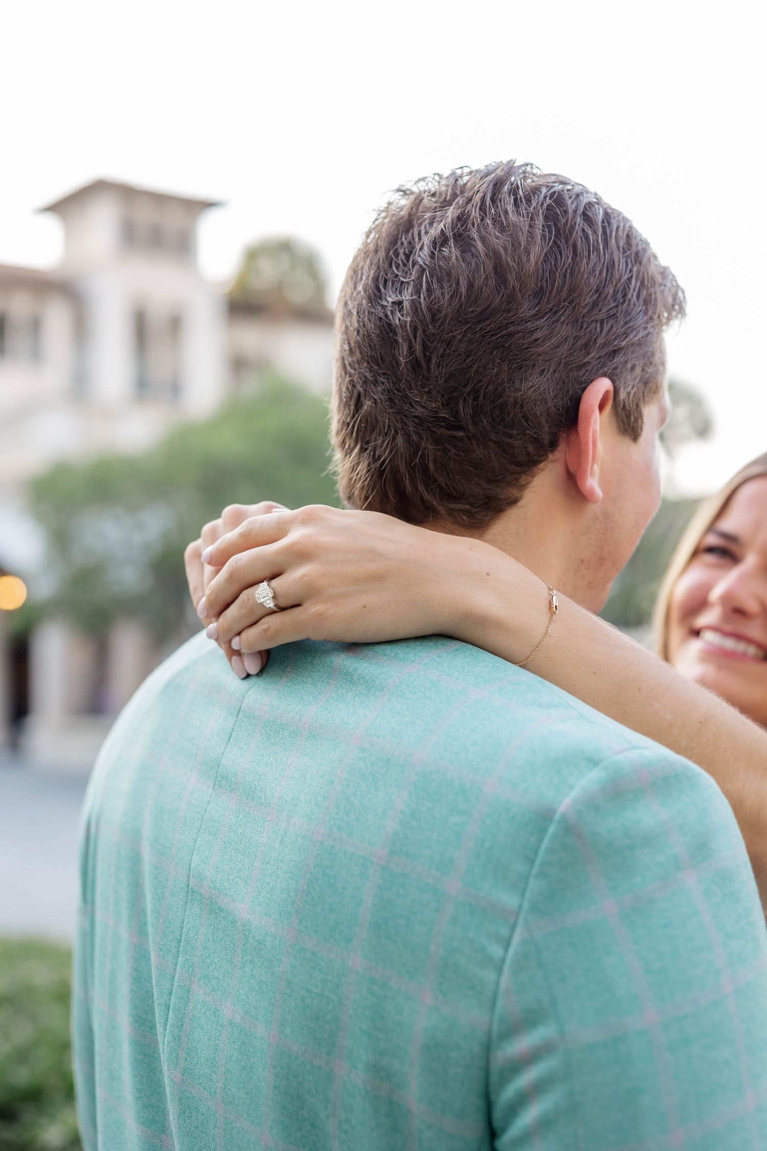 Couple embracing outdoors, woman smiling and resting hand with diamond engagement ring on man's shoulder.