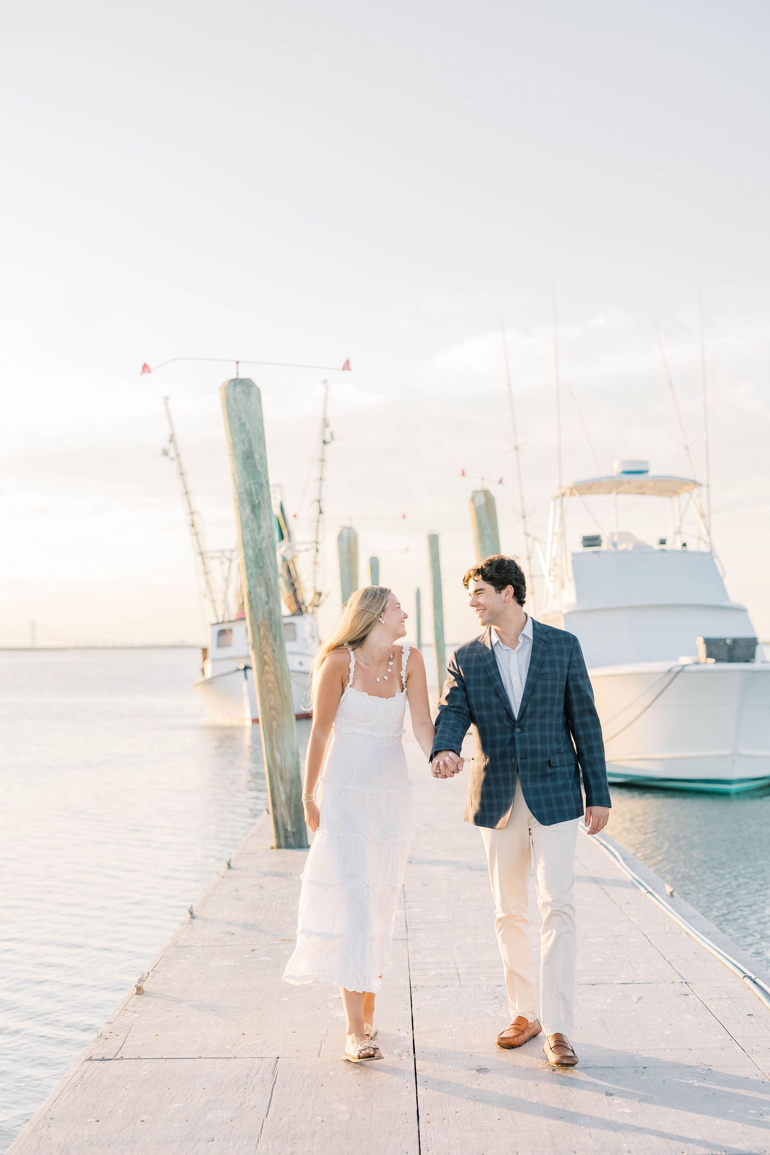 Couple holding hands walking on marina dock at sunset, woman in white dress, man in plaid blazer, boats in background.