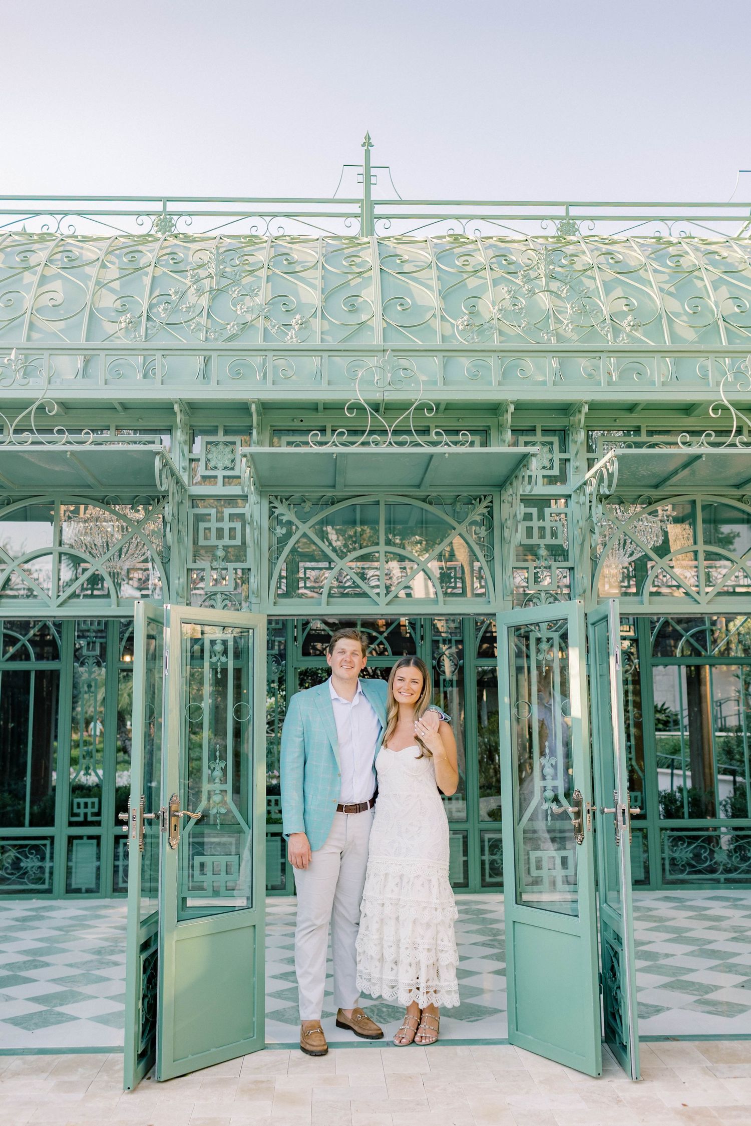 Couple posing in front of an ornate mint green iron greenhouse structure with decorative arches and open doors.
